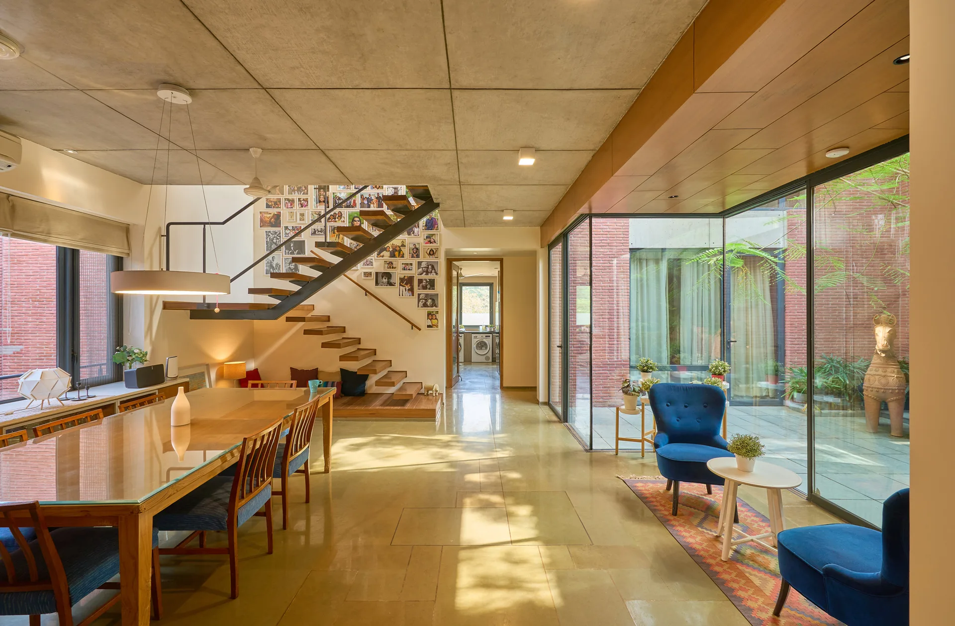 An open dining room featuring a glass-topped table, a floating wooden staircase with a photo gallery wall, and glass doors opening to a courtyard.