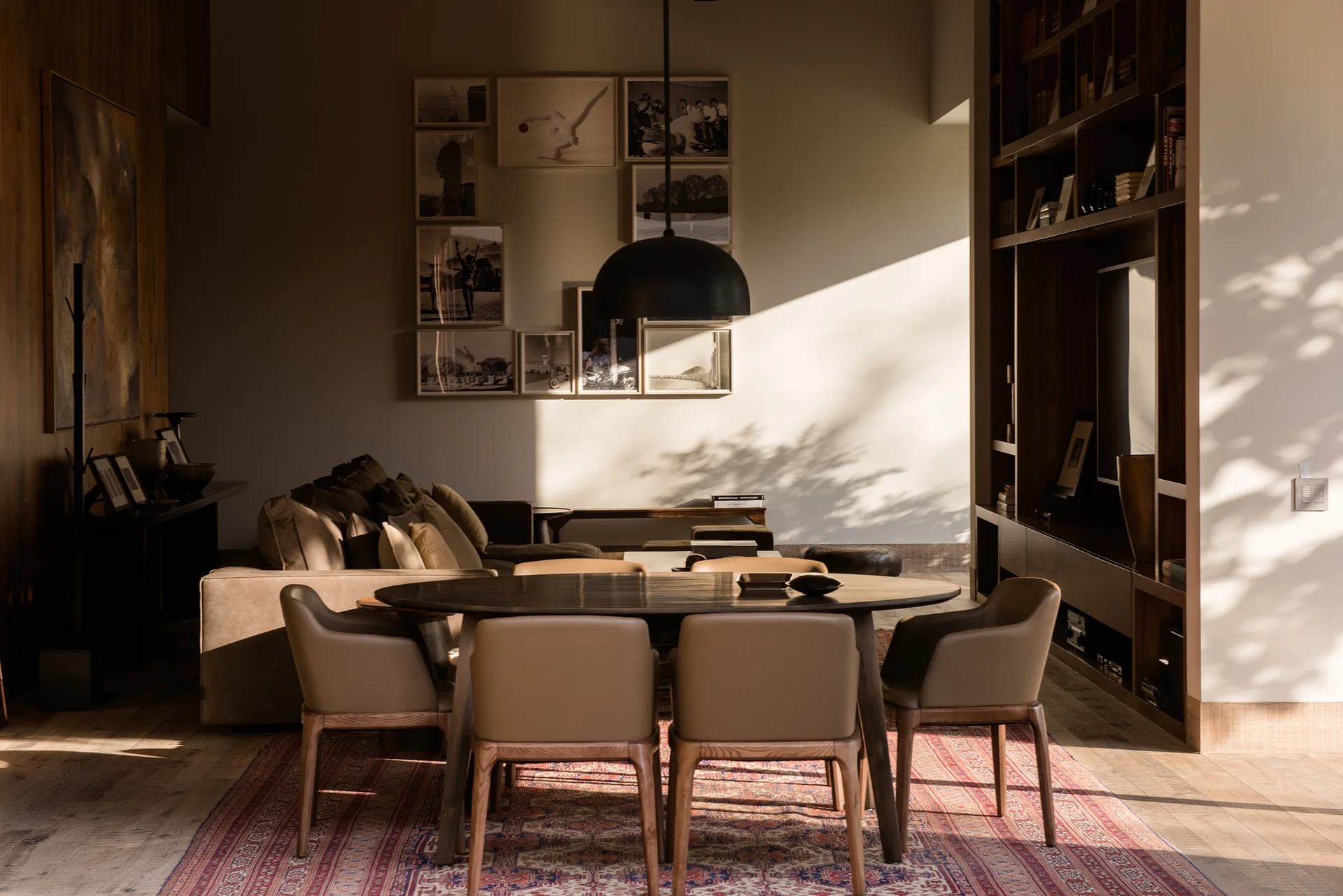 An open dining space features a dark wood oval table, tan leather chairs, a low-hanging black dome pendant light, and a patterned area rug.