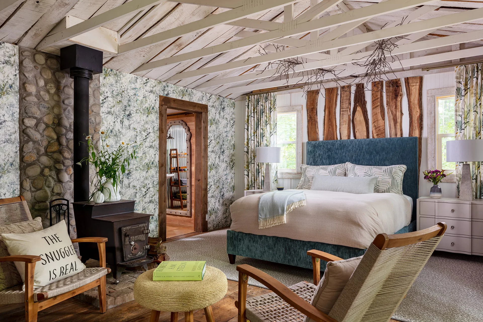 A rustic cabin bedroom features a blue upholstered bed, vertical live-edge wood panels, and a black cast-iron stove beside a stone accent wall.