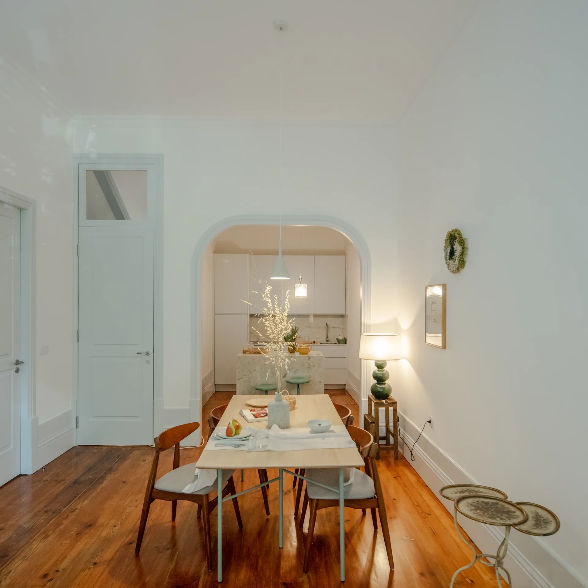 A bright dining room with warm wood floors features a light wood table under a pendant light, looking through an arched opening into a white kitchen.