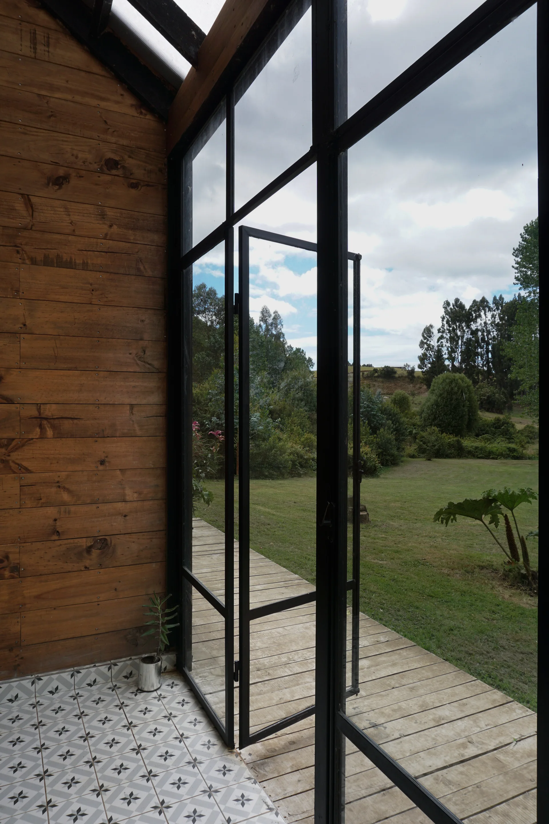 A sunroom with warm wood wall paneling and geometric floor tiles features large black metal-framed glass doors opening to an outdoor wooden deck.