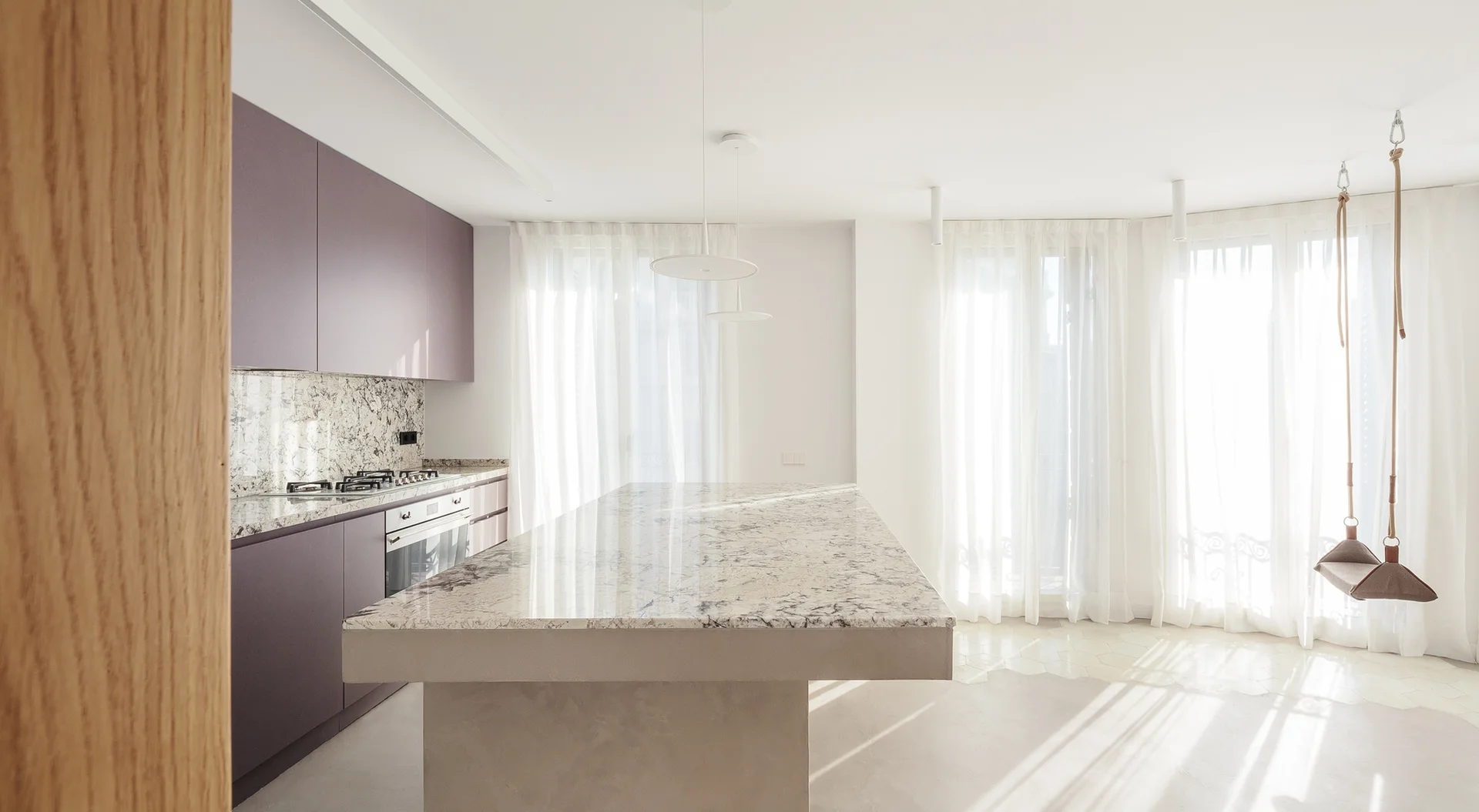 A minimalist open kitchen featuring muted mauve cabinets, a large marble kitchen island, and a hanging swing chair near sheer curtained windows.