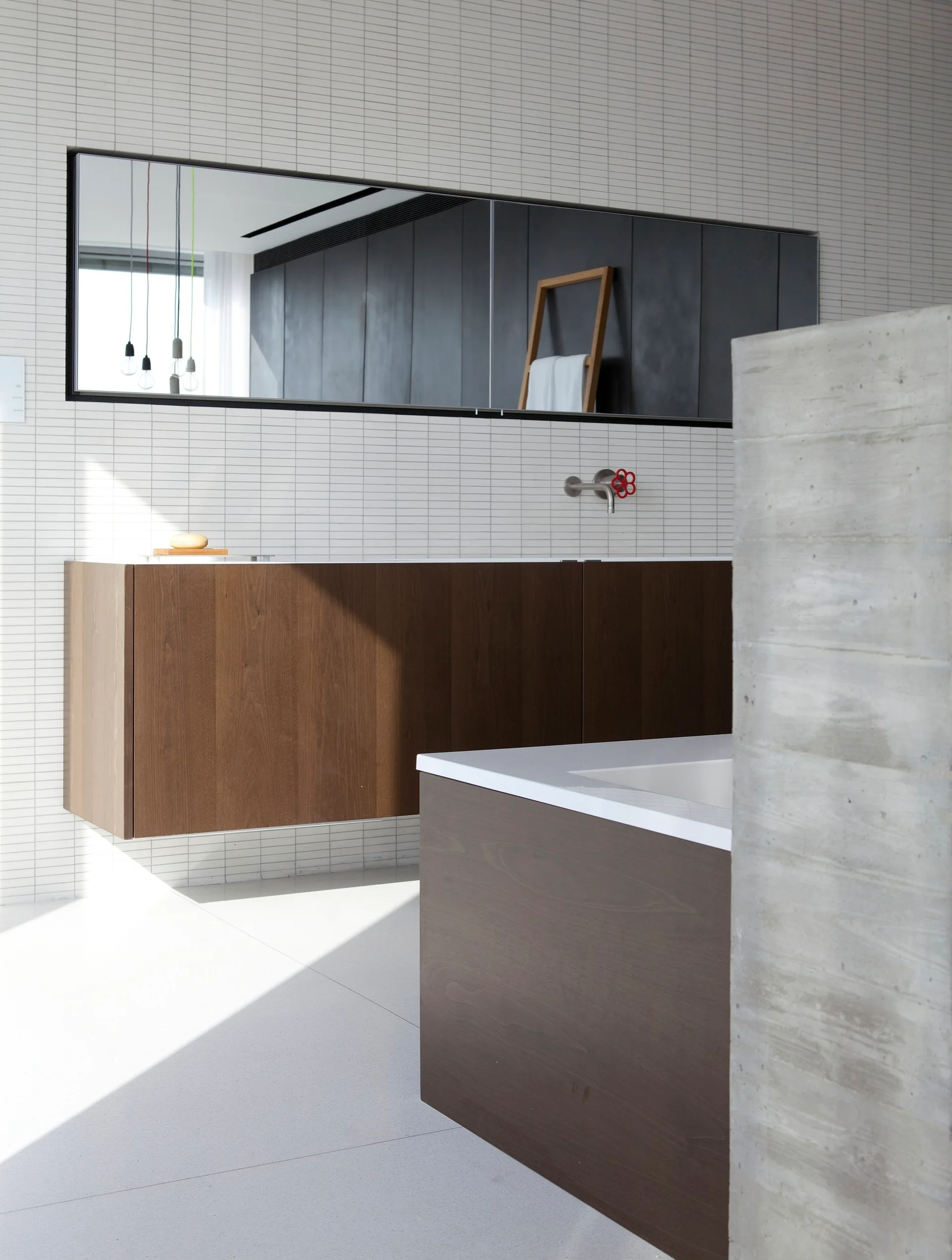 A minimalist bathroom features a wall-mounted wooden vanity, a matching wood-wrapped bathtub, and white linear tiles on the wall behind a rectangular mirror.