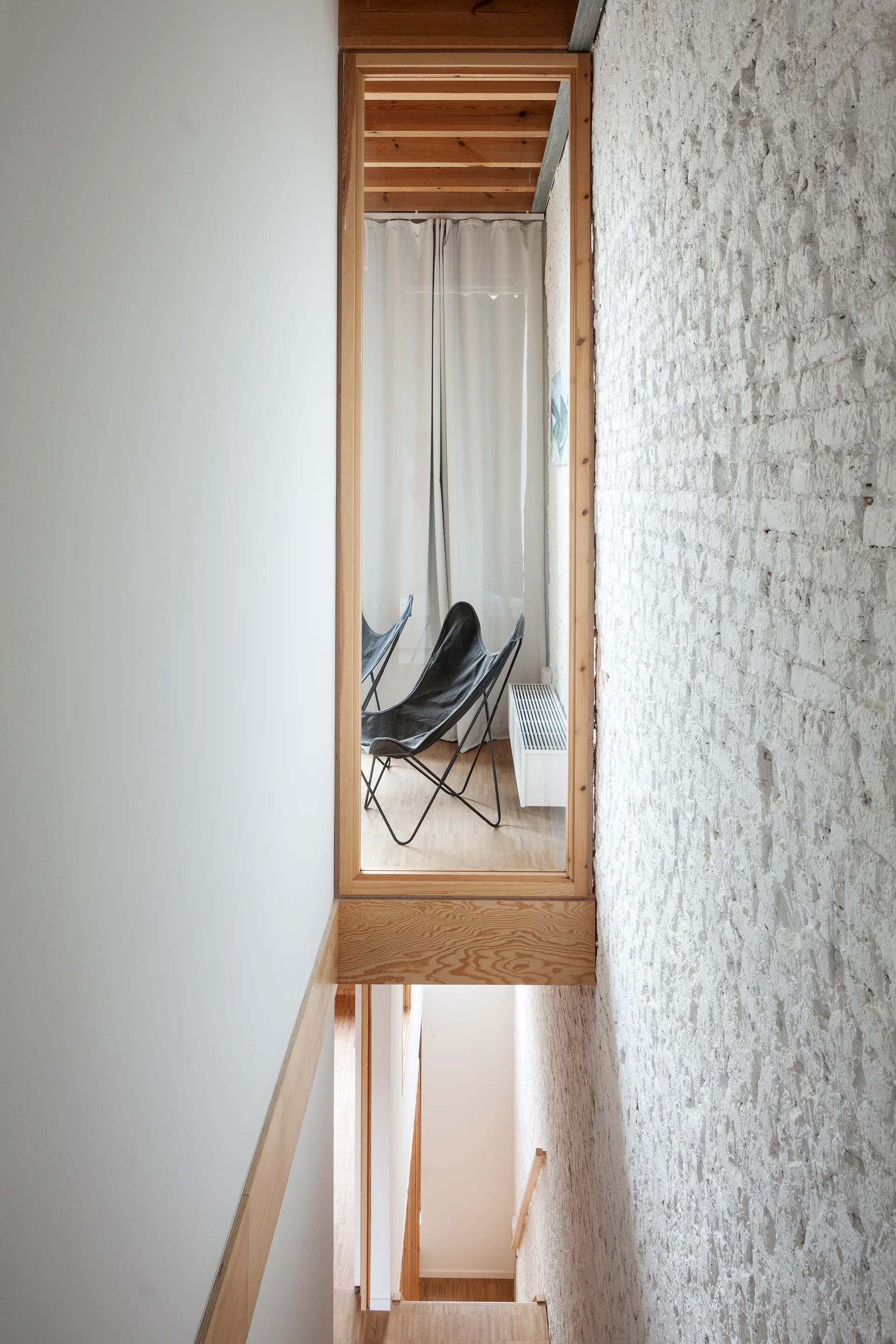 An interior window in a natural wood frame overlooks a stairwell next to a textured white painted brick wall and black butterfly chairs.