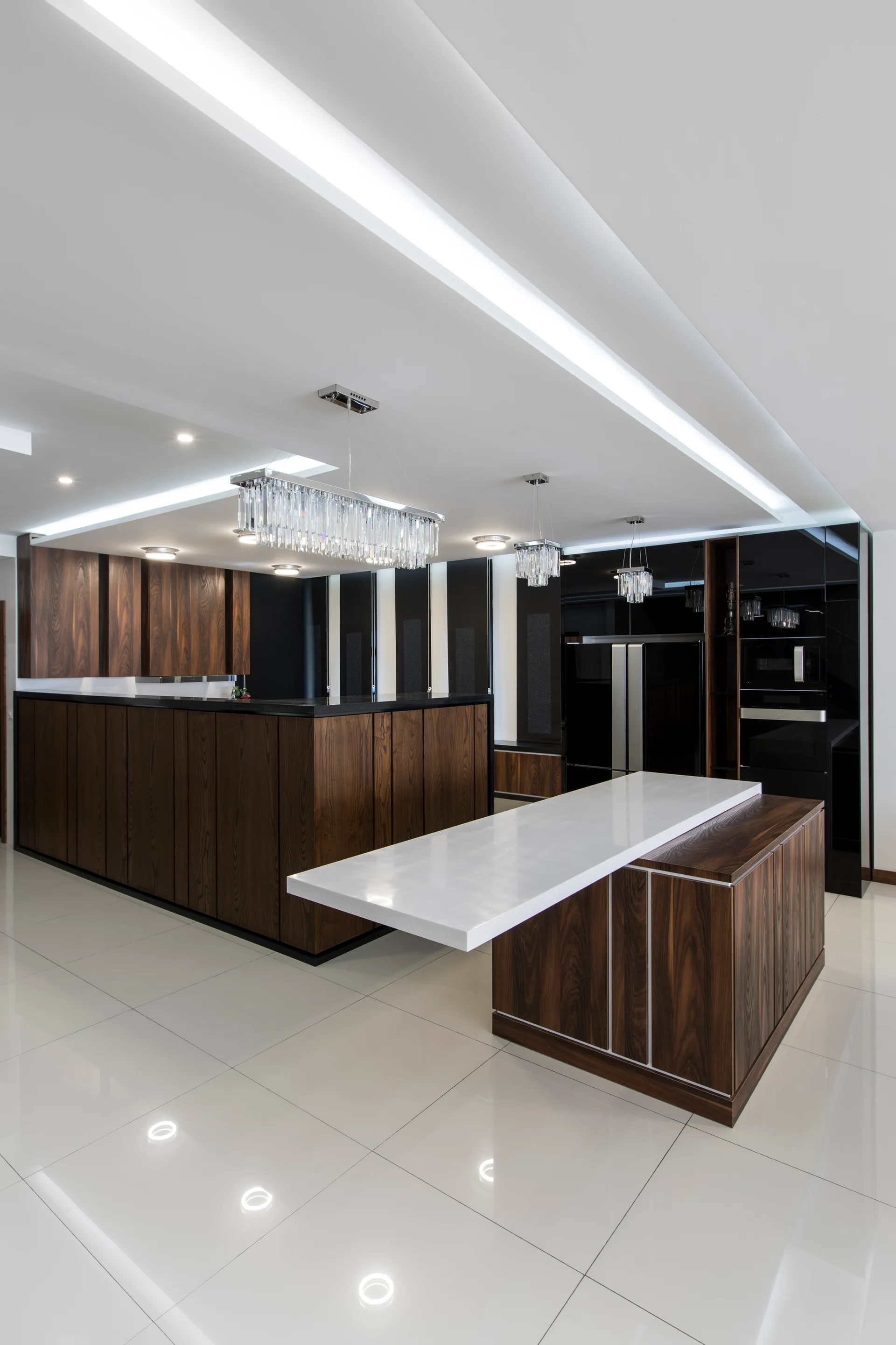 A modern kitchen features dark wood cabinetry, a two-tiered island with a white stone countertop, high-gloss white tile floors, and crystal chandeliers.