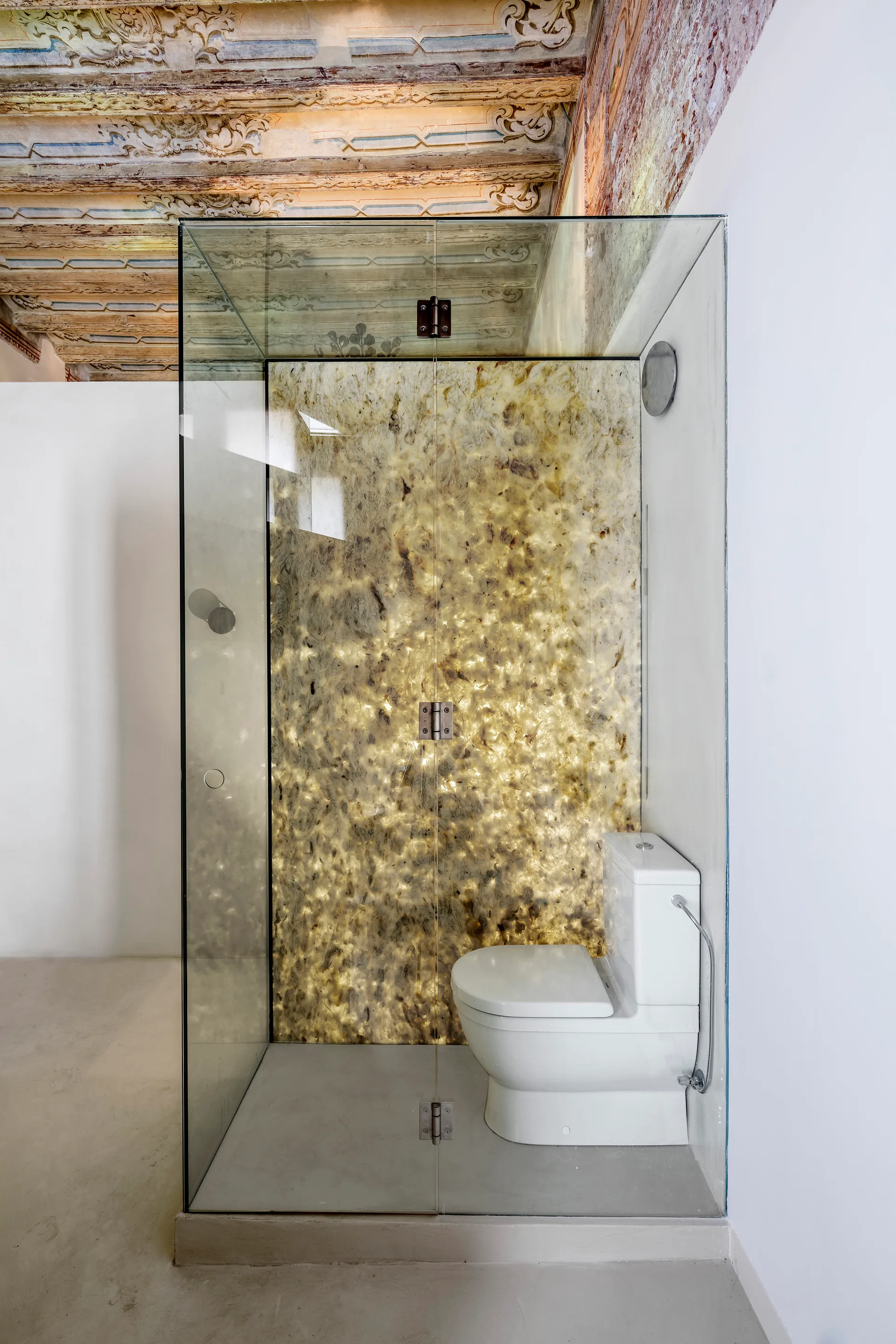 A modern glass bathroom enclosure features a white toilet set against a glowing, backlit gold stone wall beneath a decorative painted wood ceiling.