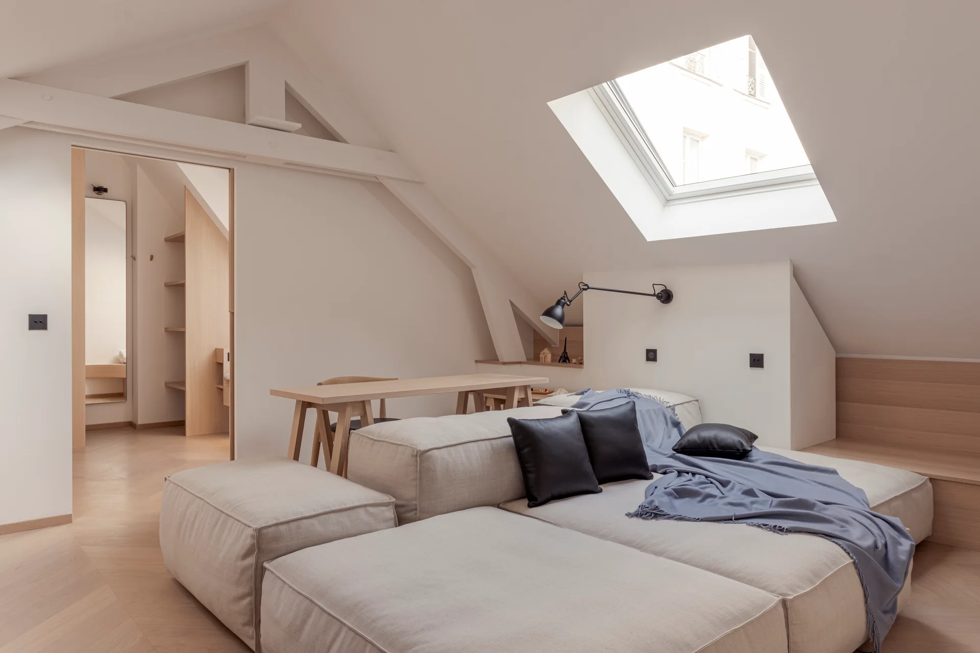 A light-filled attic bedroom features a low-profile beige modular sofa, a wooden trestle table, a black wall lamp, and a skylight on a sloped ceiling.