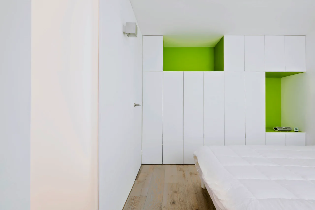 A minimalist bedroom featuring a built-in white wardrobe with lime green open shelving next to a white bed.