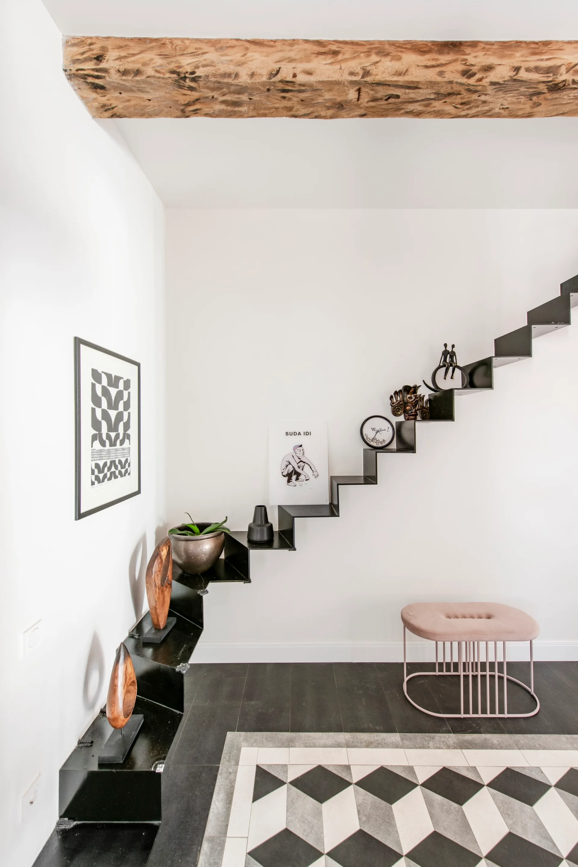 A black metal floating staircase decorated with small sculptures sits against a white wall above a geometric black-and-white tiled floor.