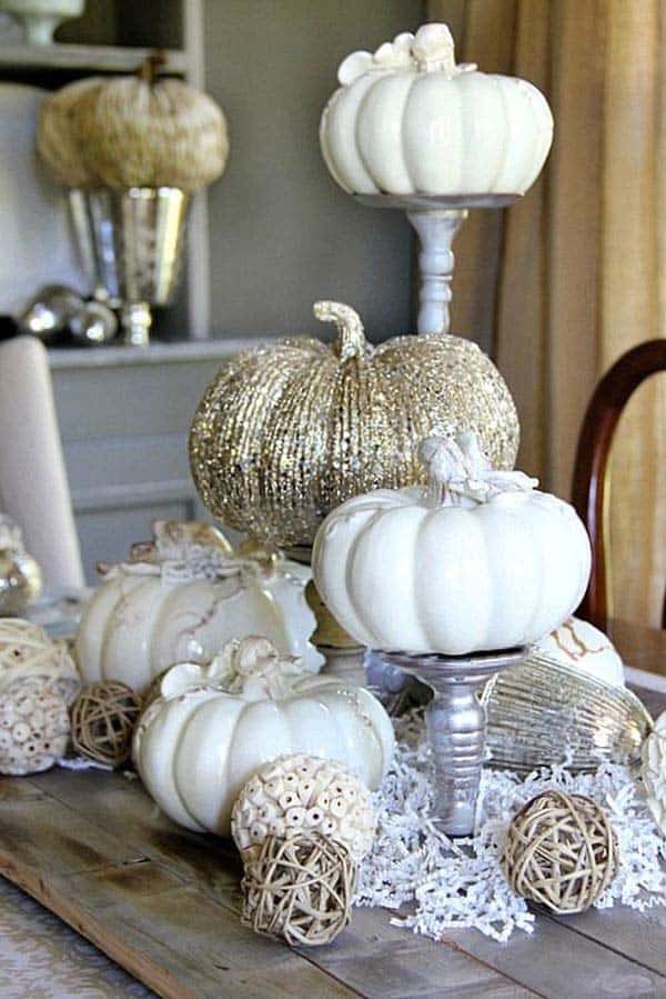 A rustic wood dining table features a centerpiece of white and metallic pumpkins displayed on silver pedestals alongside textured woven spheres.