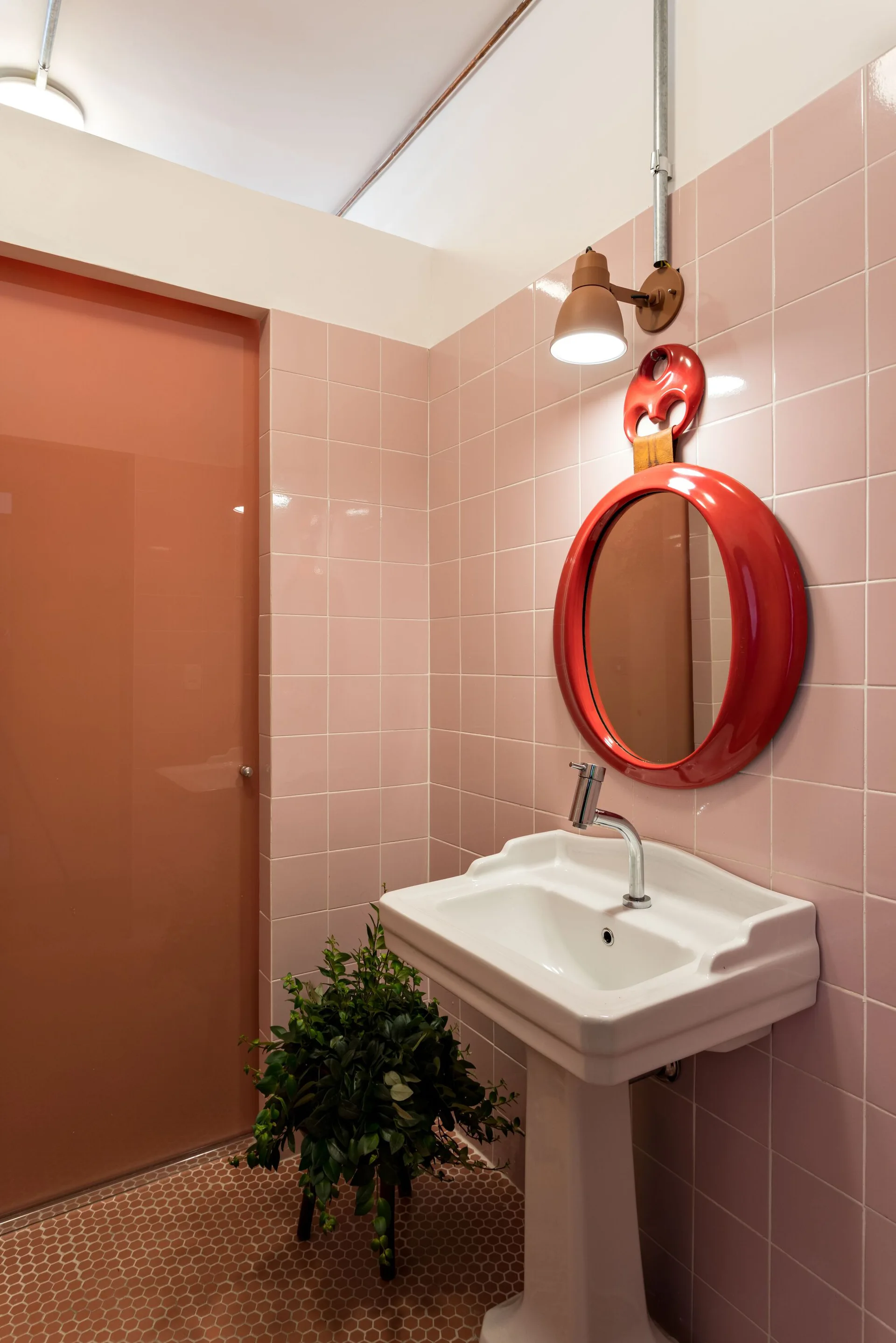 A bathroom featuring pink square wall tiles, a white pedestal sink, a glossy red circular mirror, and hexagonal floor tiles.