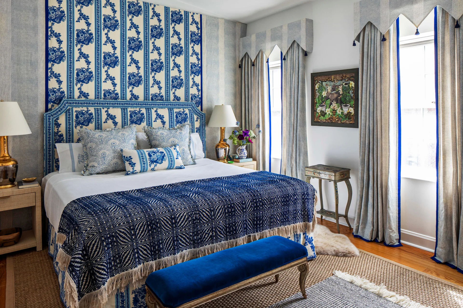 A traditional bedroom features a blue and white toile patterned headboard, matching wall hanging, patterned curtains, and a blue velvet bench.