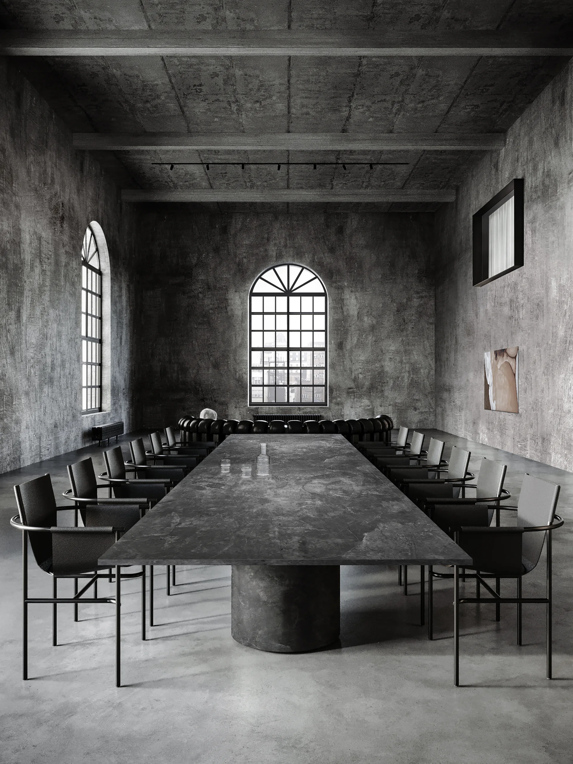 A long stone dining table and dark minimalist chairs sit inside a spacious industrial room with textured concrete walls and large arched windows.
