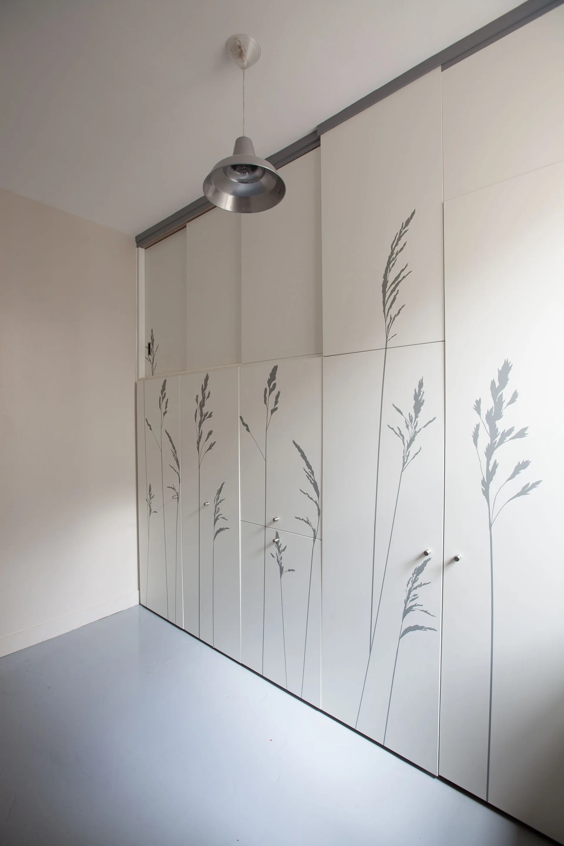 A minimalist white built-in wardrobe features a tall grey botanical grass graphic stenciled across its flat panel doors.