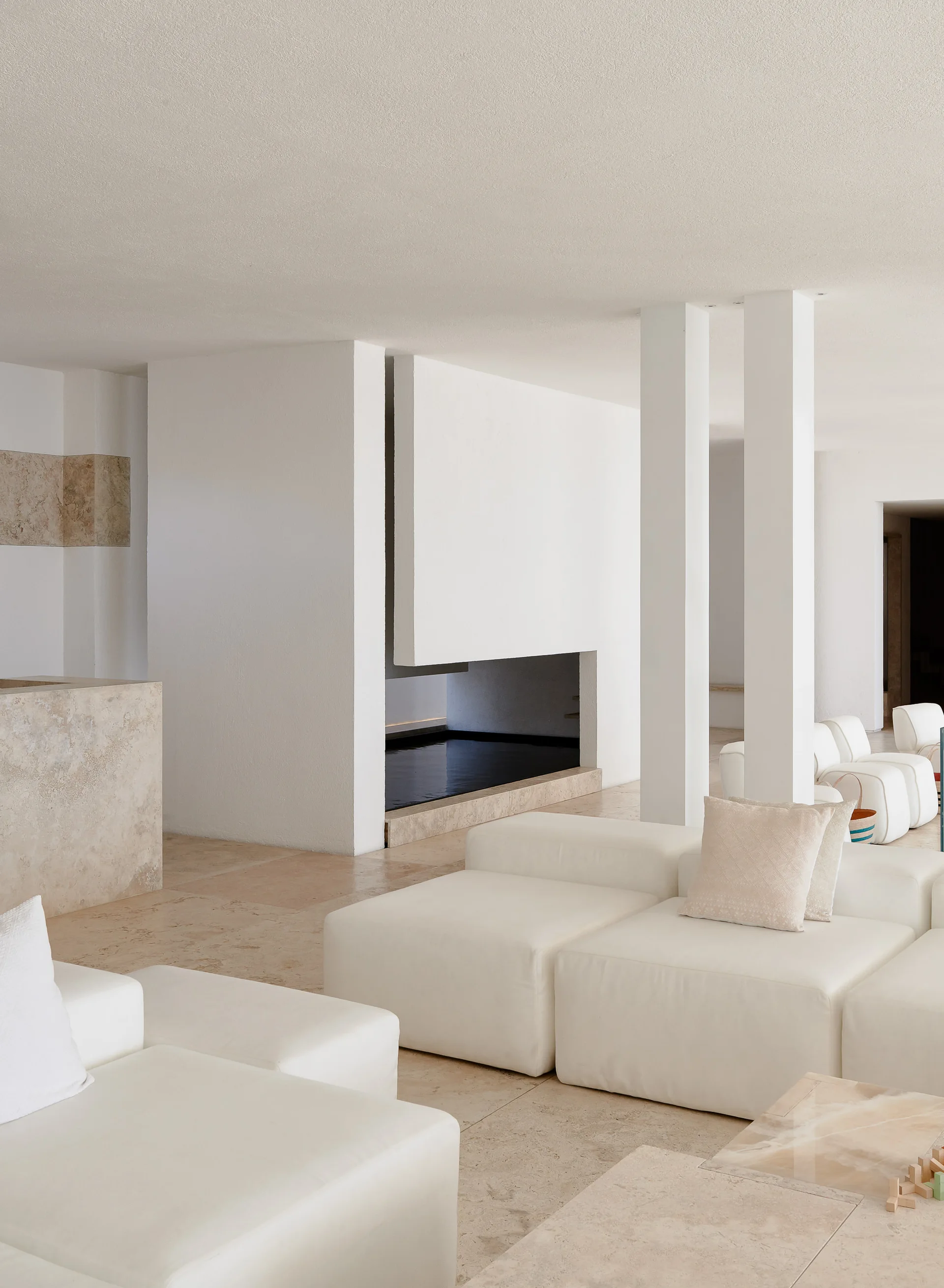 A spacious minimalist living room features white modular low-profile sofas on limestone tile flooring near an architectural white fireplace.