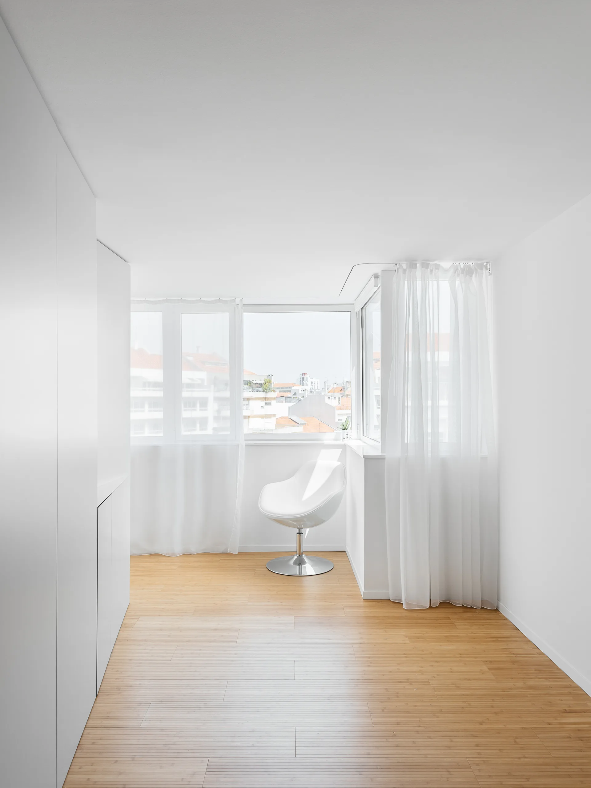 A minimalist white room with light wood flooring features a single white modern pedestal chair next to a corner window with sheer curtains.