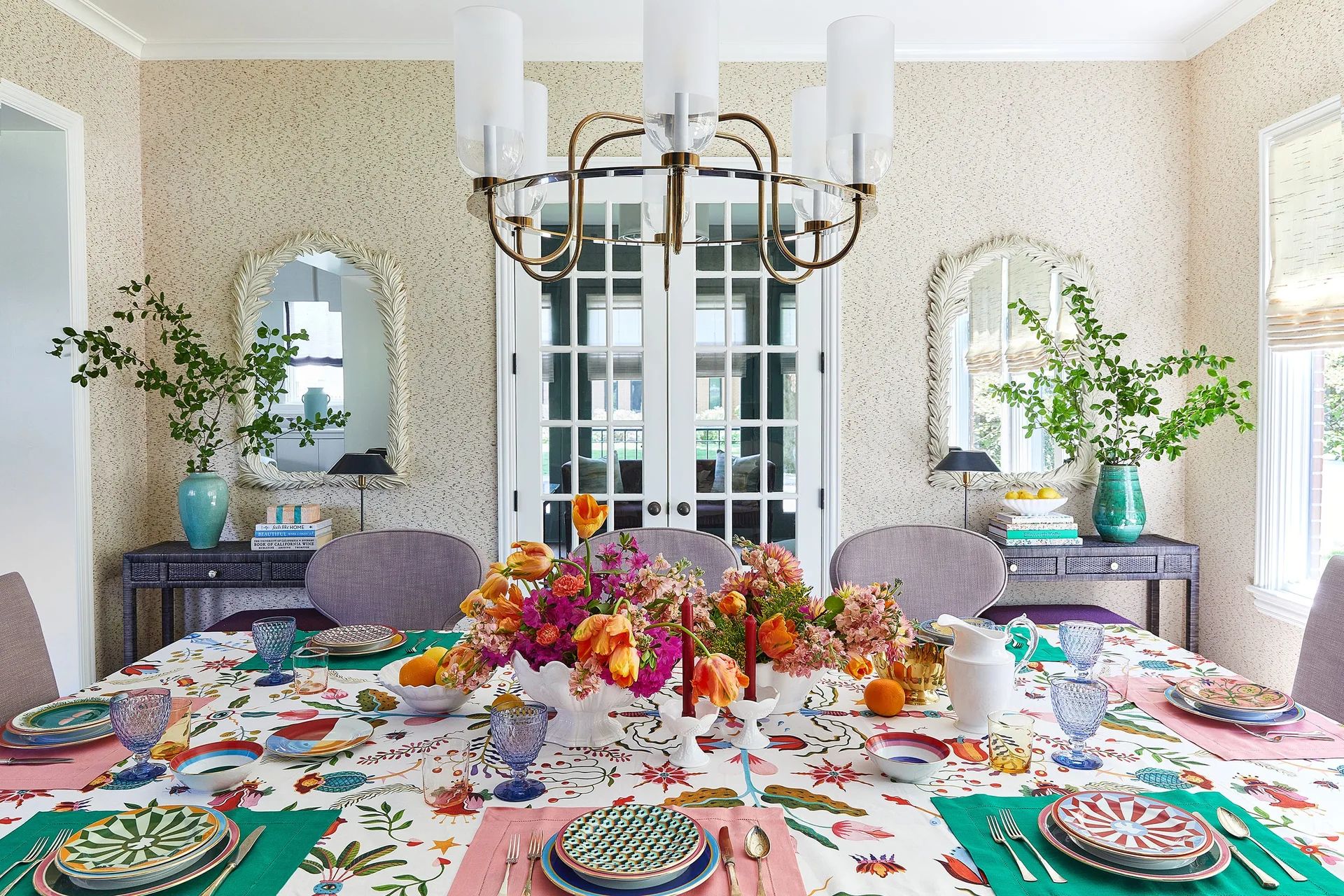 A dining table set with a floral tablecloth, colorful tableware, and a vibrant flower centerpiece in front of white French doors.