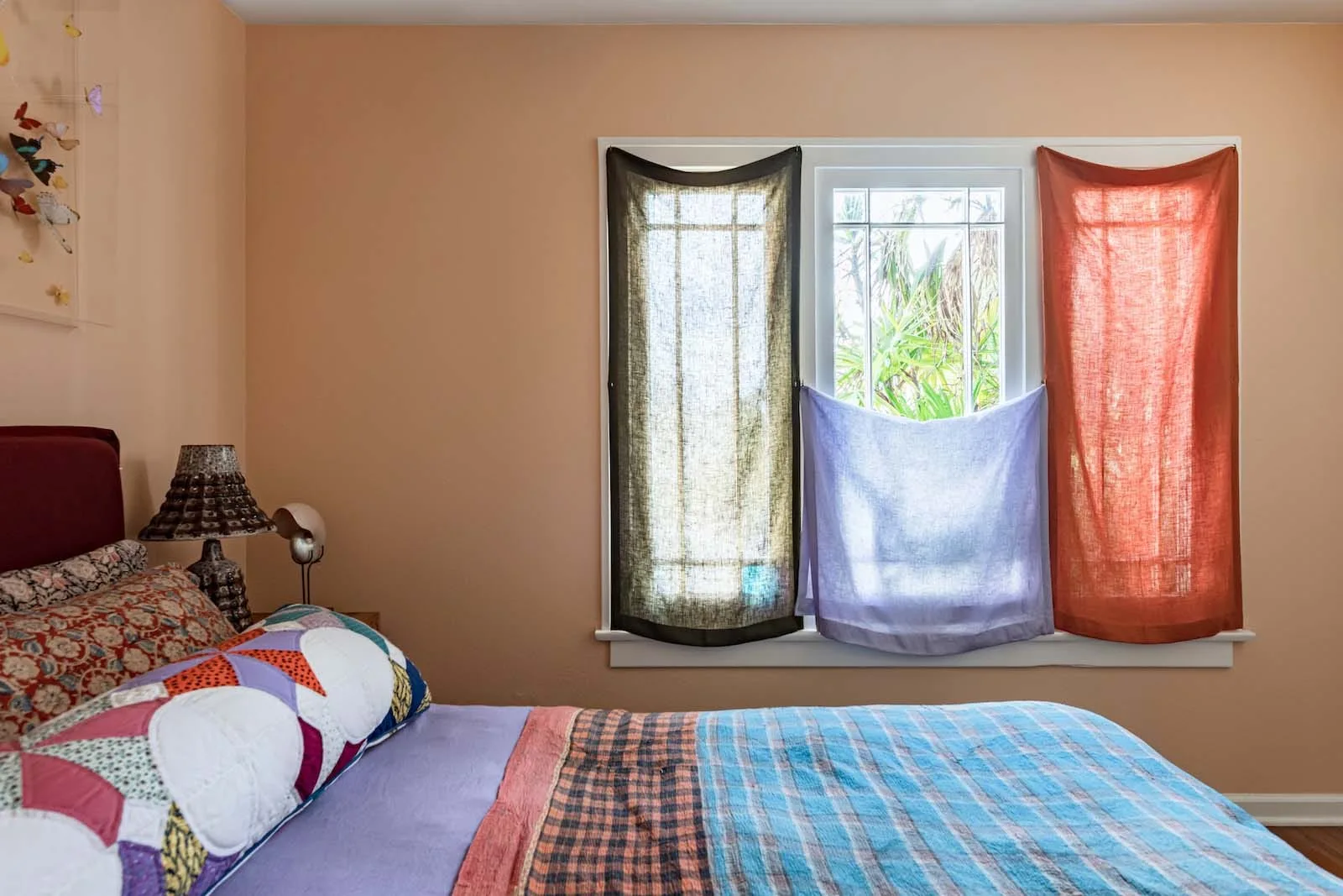 A bedroom featuring peach walls, a bed with a colorful patchwork quilt, and a window styled with three draped translucent fabric panels.
