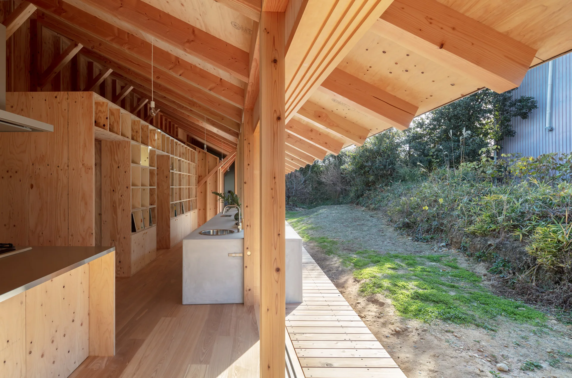 A minimalist kitchen features exposed plywood walls, a concrete island with a sink, and a sloped ceiling opening to an outdoor deck.