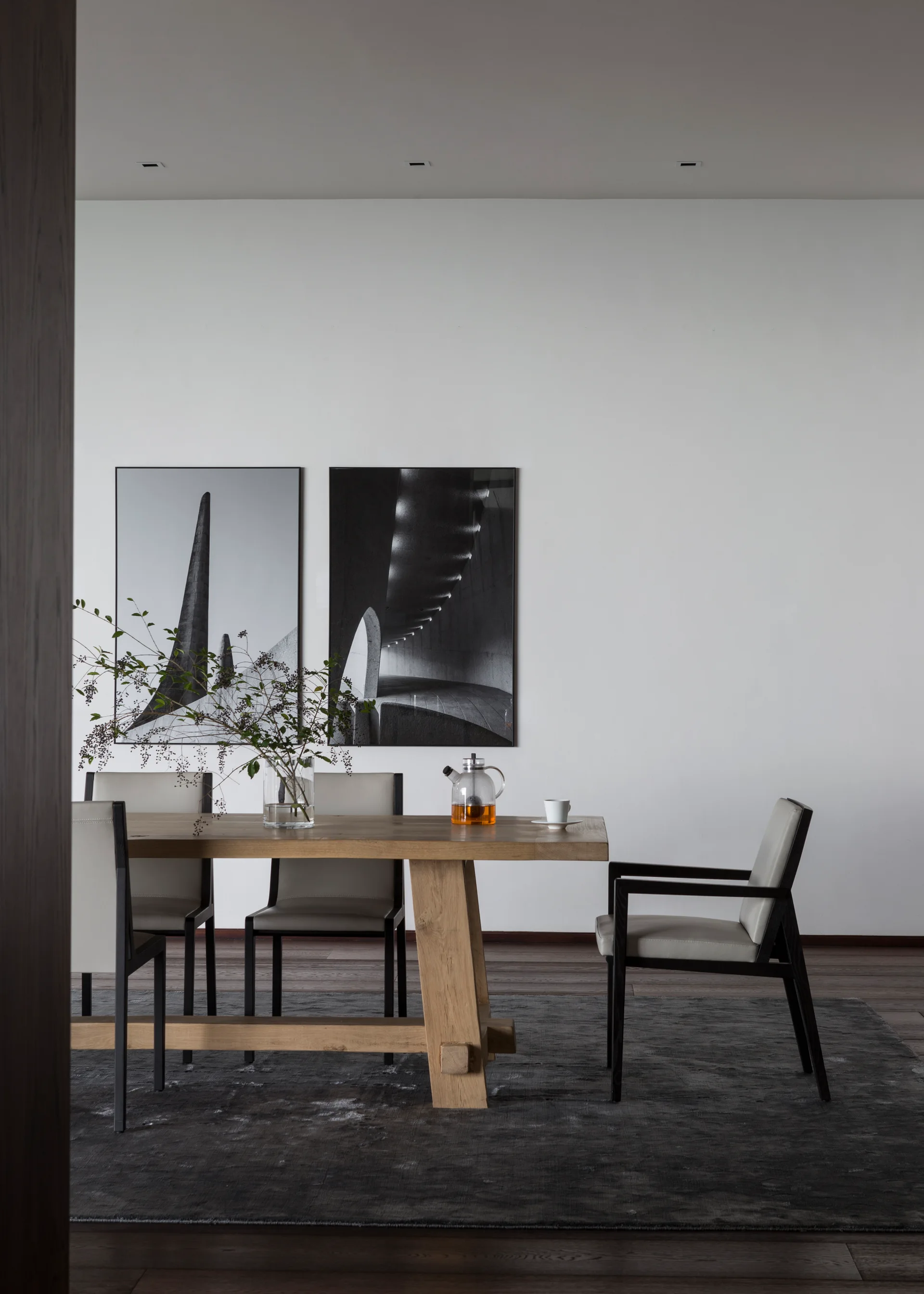 A light wood dining table with upholstered chairs sits on a dark gray rug in front of a white wall decorated with two framed black and white photos.
