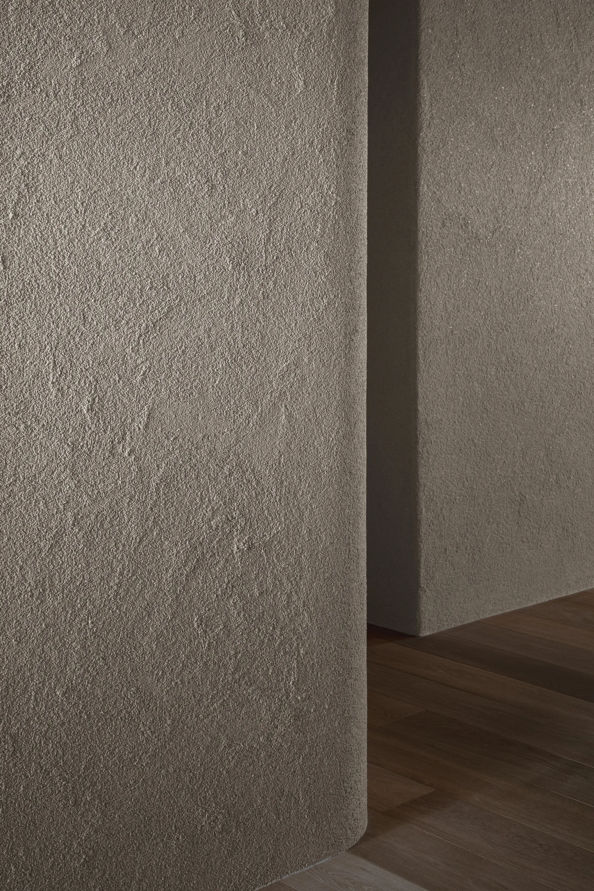 A close-up of a textured greige plaster wall with a curved corner meeting dark hardwood flooring.