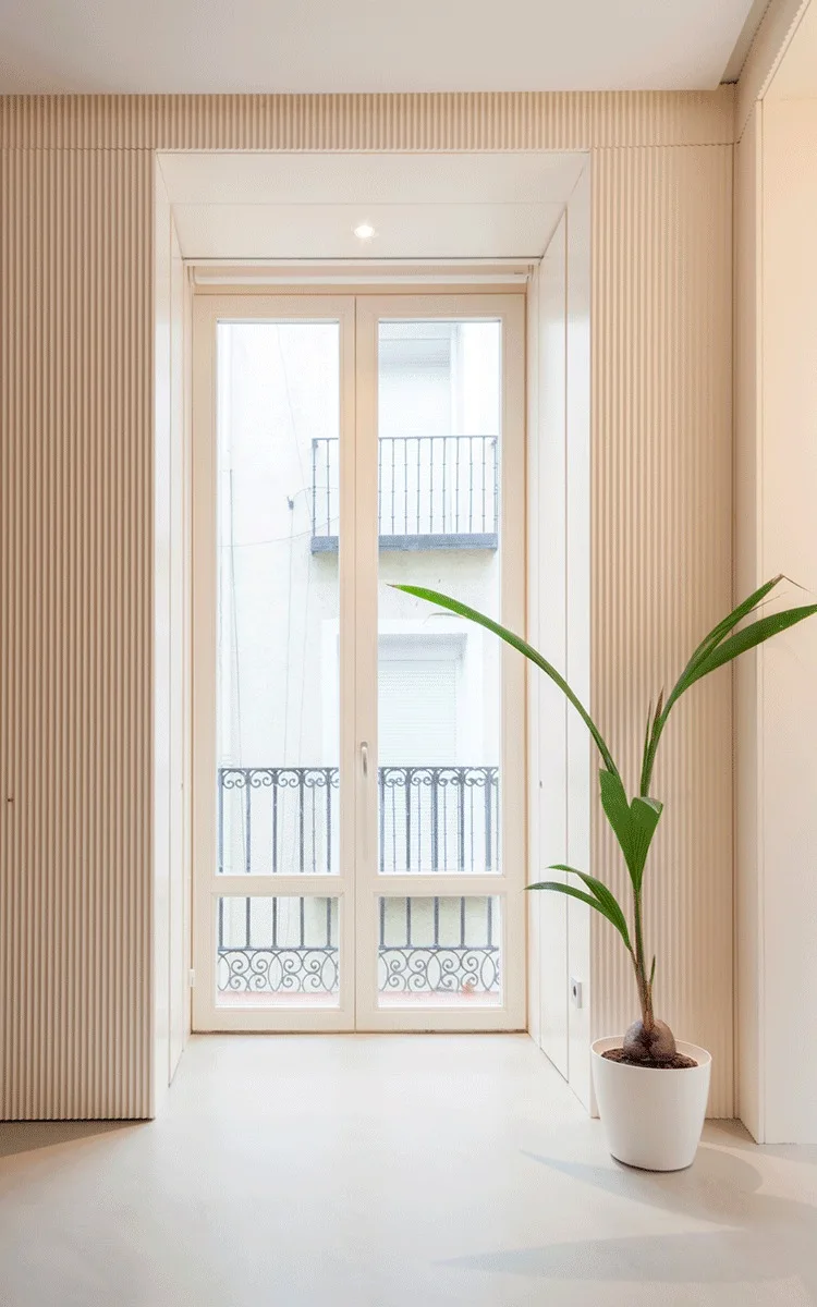 A minimalist room with cream fluted wall panels framing a tall glass French door that opens onto a balcony with black metal railings.