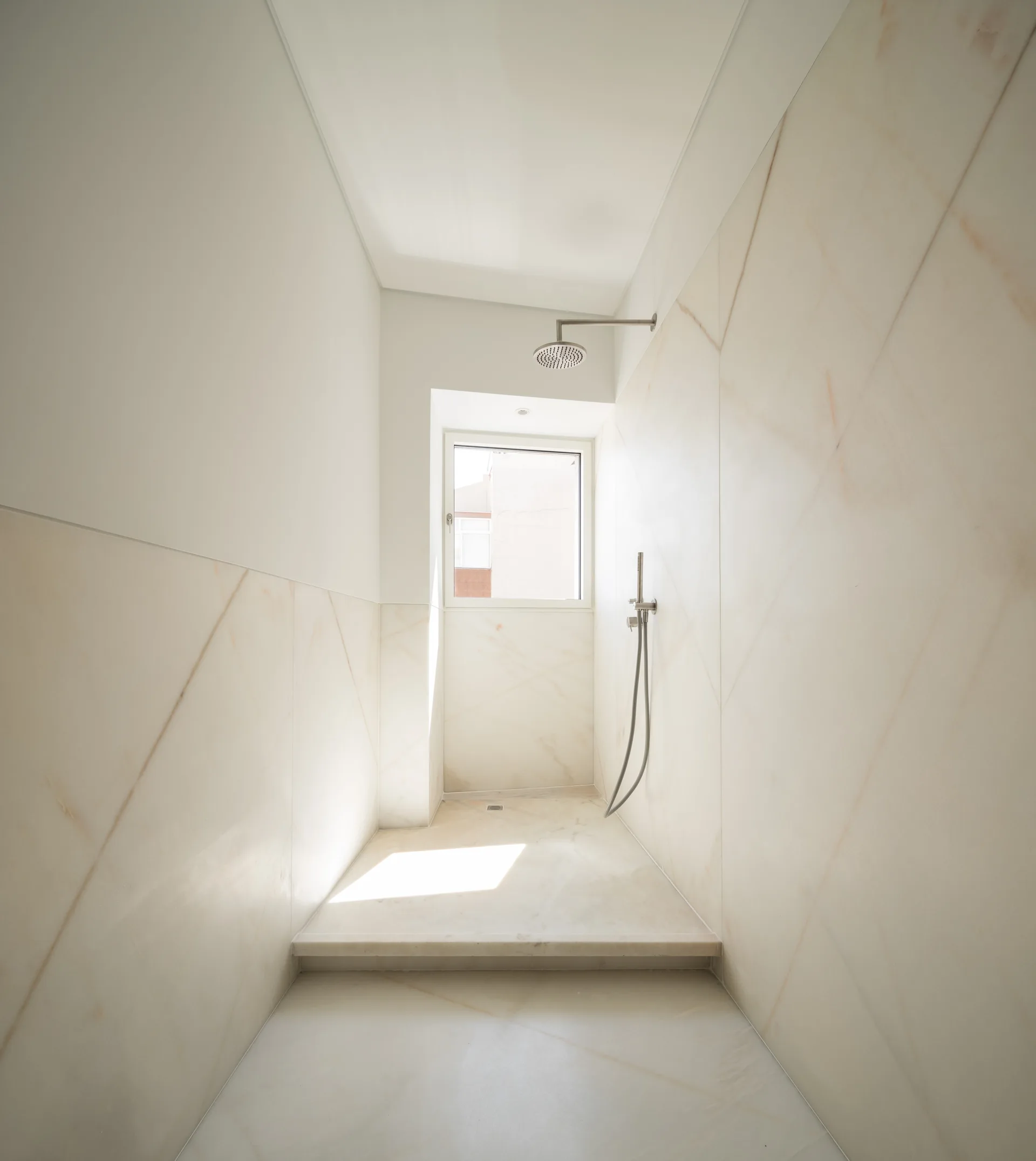 A minimalist walk-in shower features large-format cream veined tiles, a rainfall shower head, and a small square window letting in natural light.