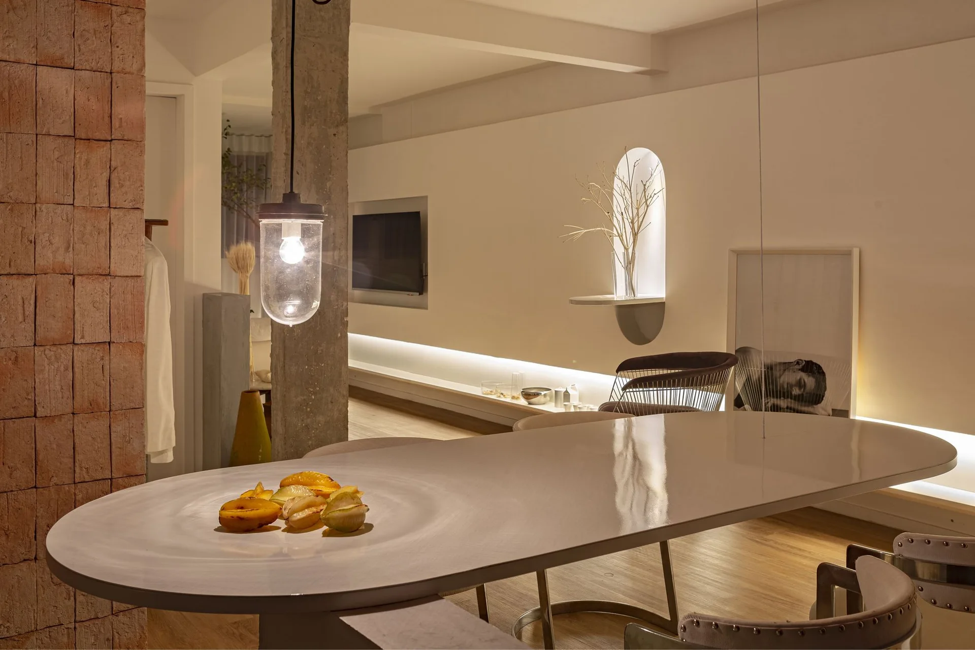 A modern dining area features a glossy oval table, an exposed concrete pillar, a glass pendant light, and an illuminated arched wall niche in the background.