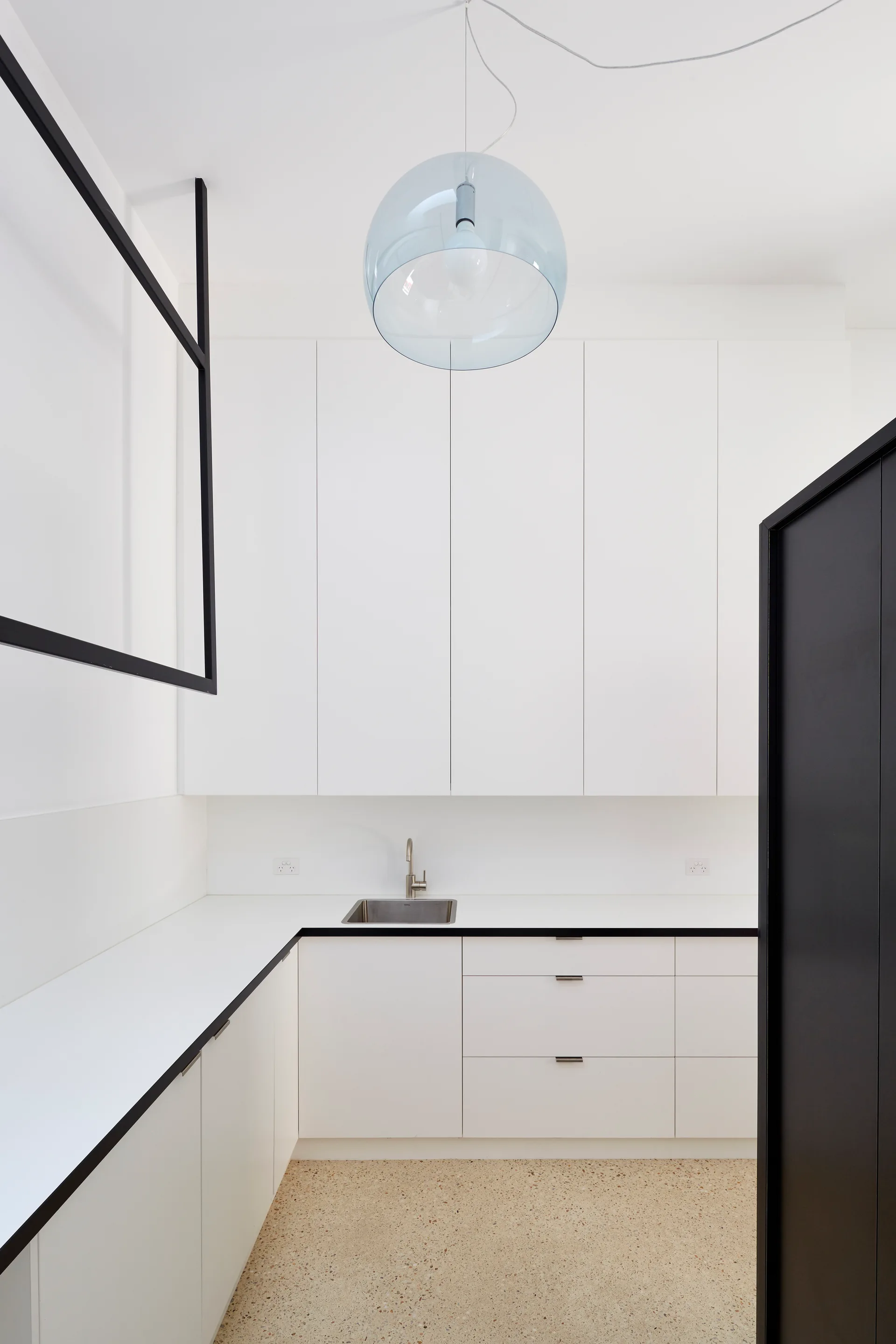 A minimalist kitchen features seamless white cabinets, a light beige terrazzo floor, a stainless steel sink, and a translucent blue pendant light.