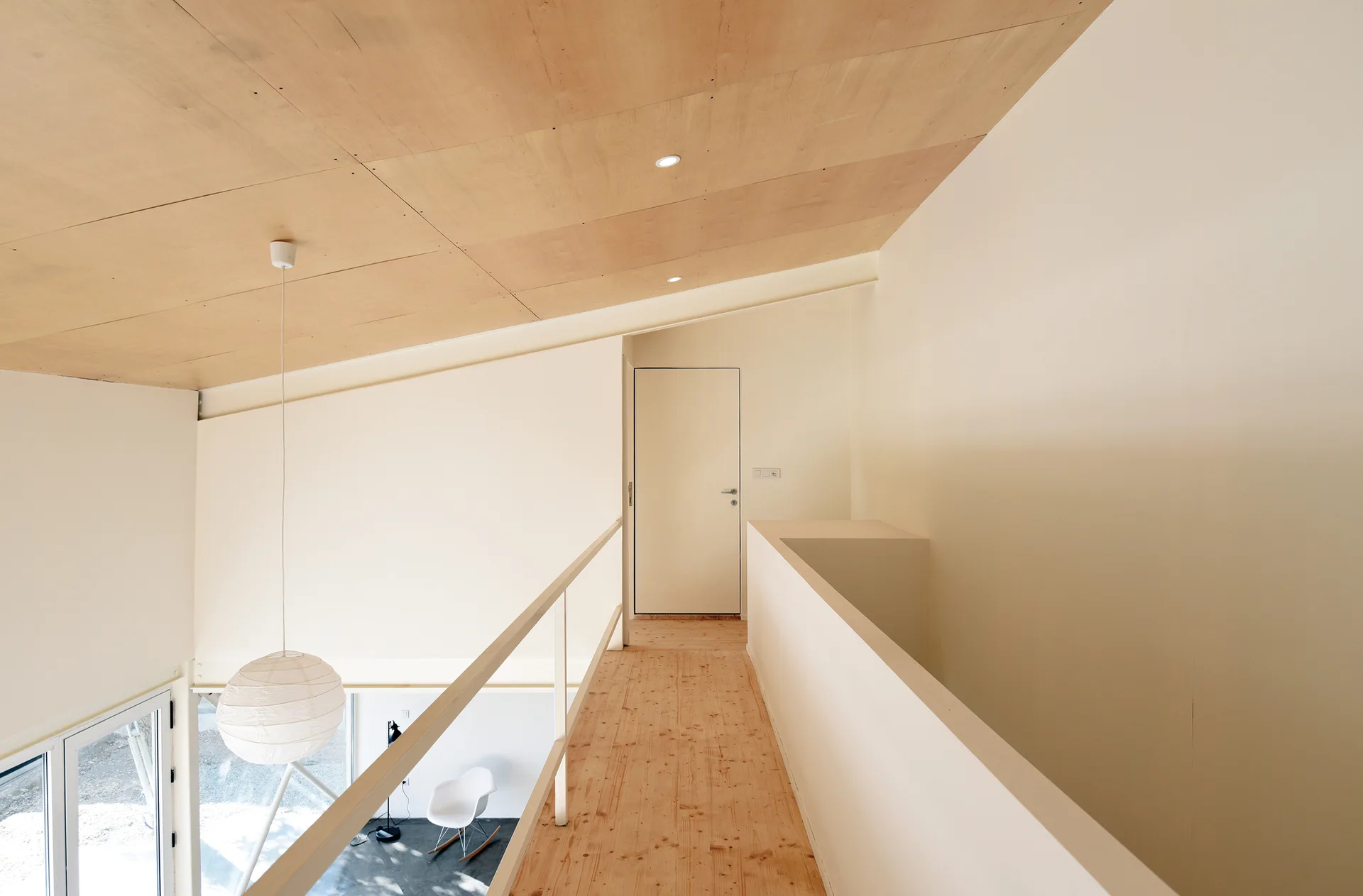 A minimalist mezzanine walkway featuring a light wood floor, a sloped plywood ceiling, off-white walls, and a simple metal railing.