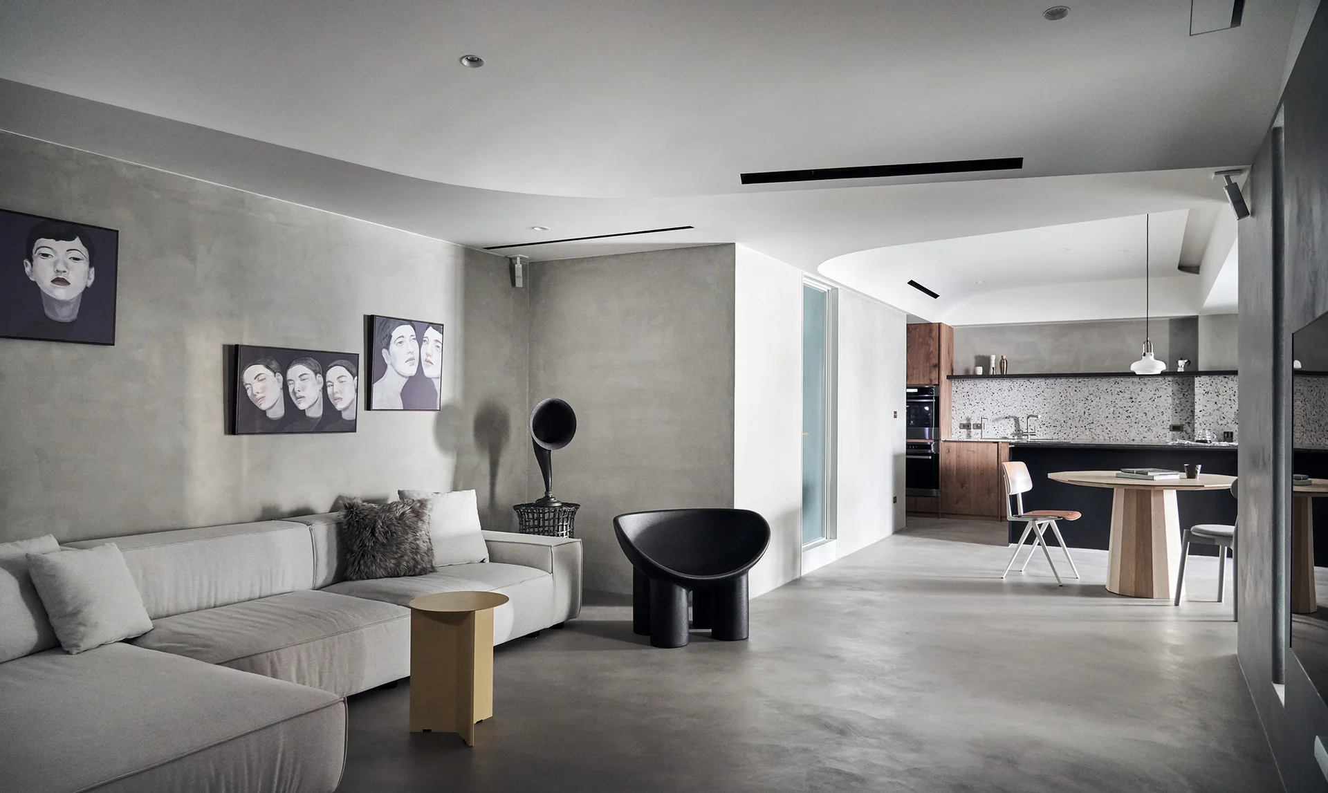 A minimalist open-plan living room features a gray sectional sofa, concrete walls, a sculptural black chair, and a curved ceiling design.