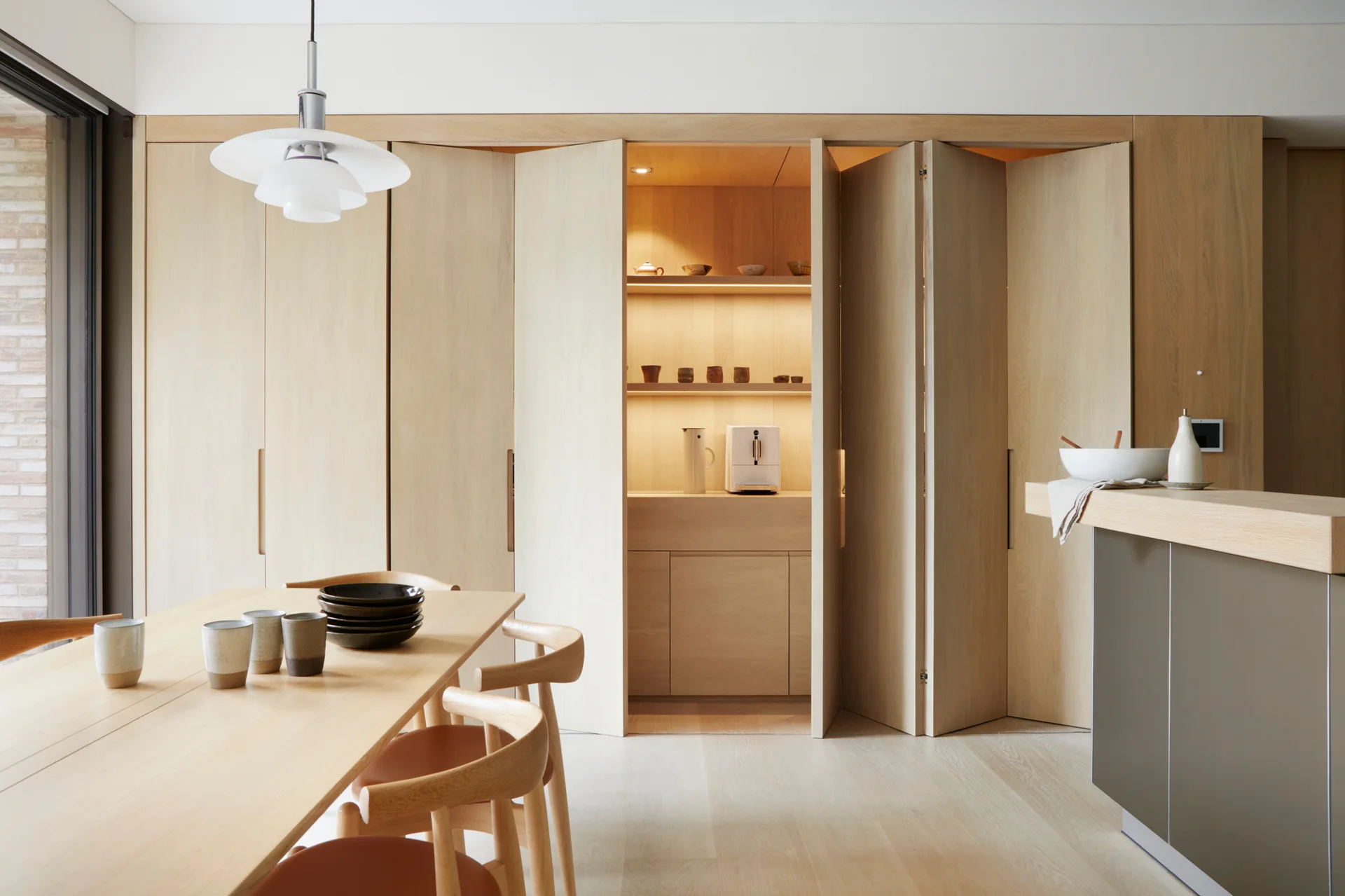 A dining room featuring a wooden table, chairs, and a wall of light wood bi-fold doors opened to reveal a lit recessed pantry shelf.