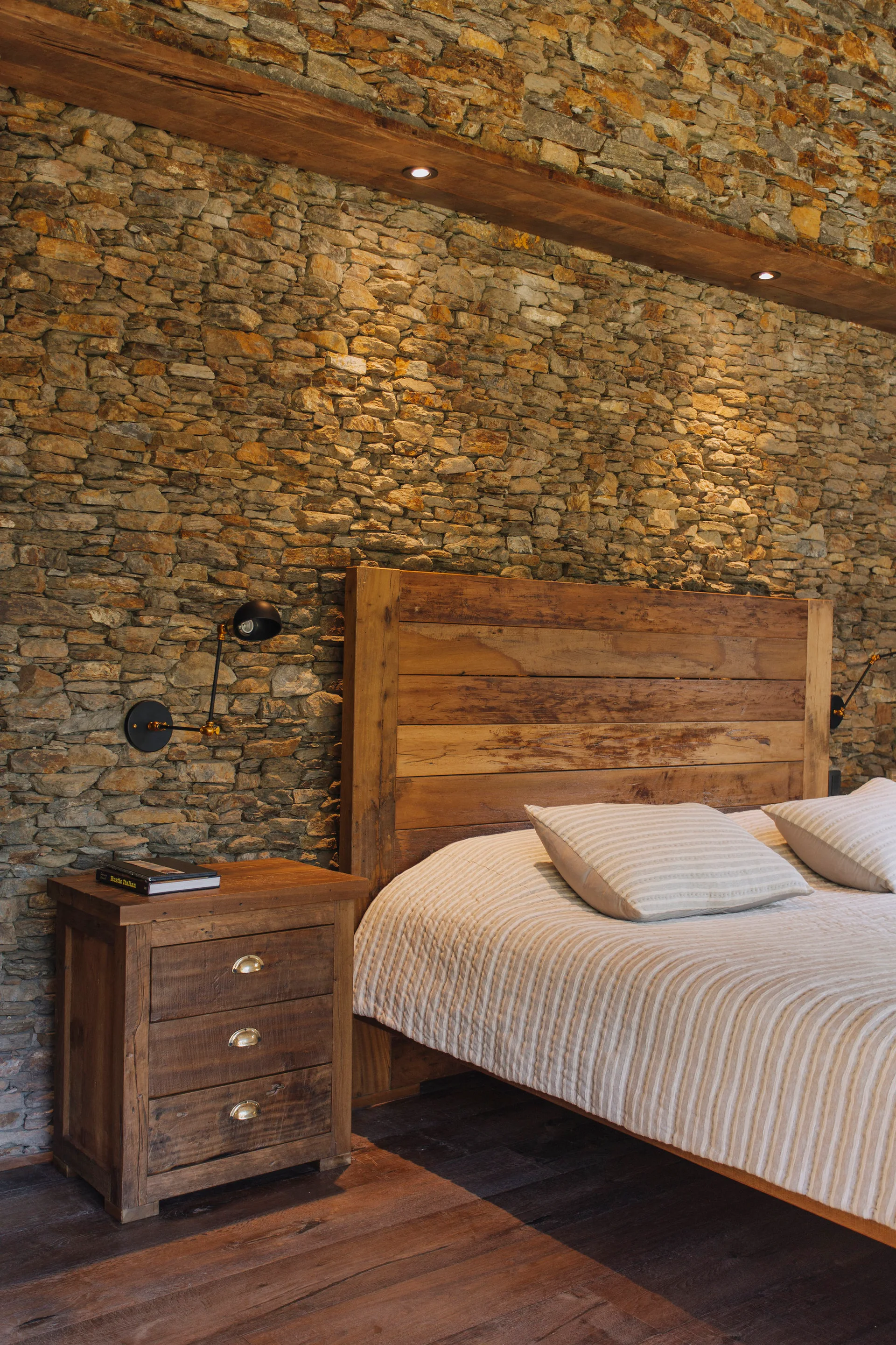 A rustic bedroom features a natural wood bed with striped bedding set against a textured stone wall alongside a matching wooden nightstand.