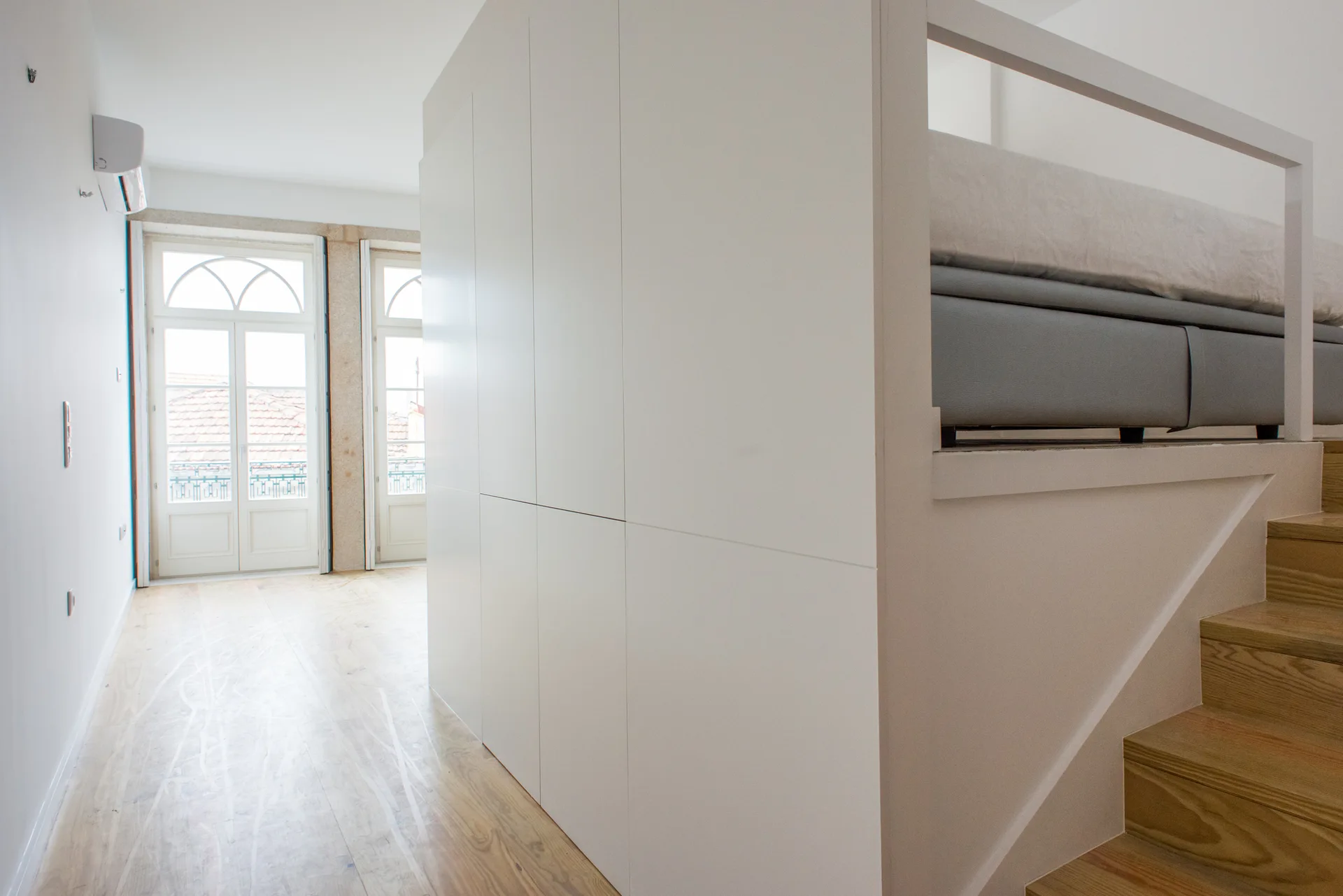 An elevated bed platform sits atop a large white built-in storage cabinet next to wooden stairs in a bright, minimalist room.