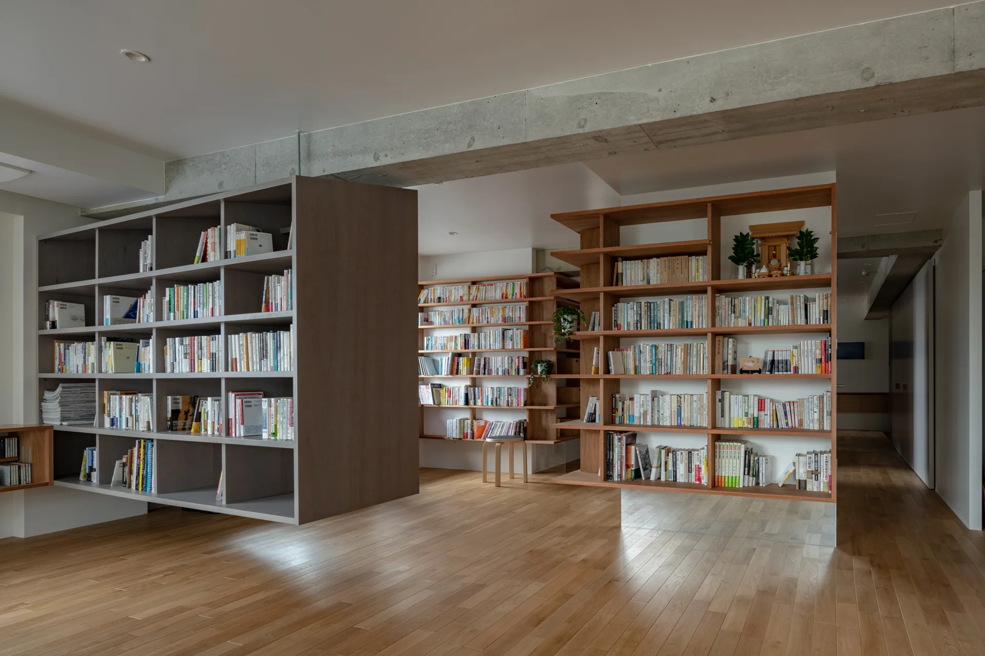 A spacious minimalist library features suspended grey and wood bookcases, light hardwood flooring, and an exposed concrete ceiling beam.