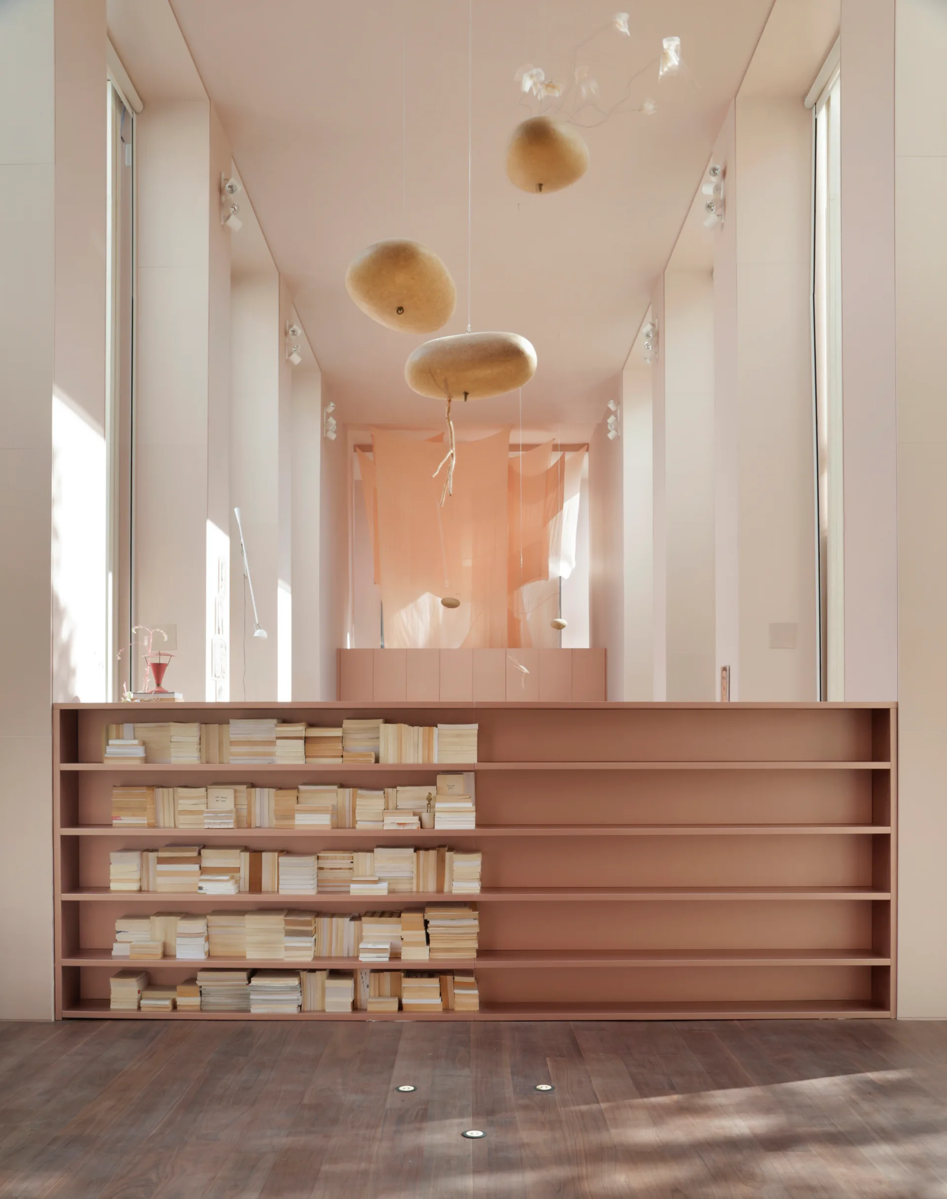 A long monochromatic peach corridor featuring a low built-in bookcase filled with books, organic-shaped pendant lights, and dark wood flooring.