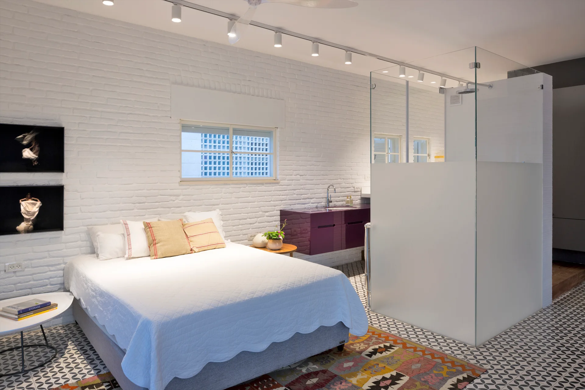 A modern bedroom featuring a bed with a white quilt, a white painted brick wall, a glass shower enclosure, and a purple floating vanity.