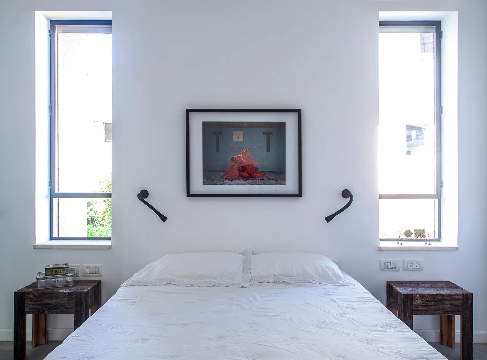 A centered white bed flanked by two narrow vertical windows, black wall sconces, and rustic wooden nightstands on a grey floor.