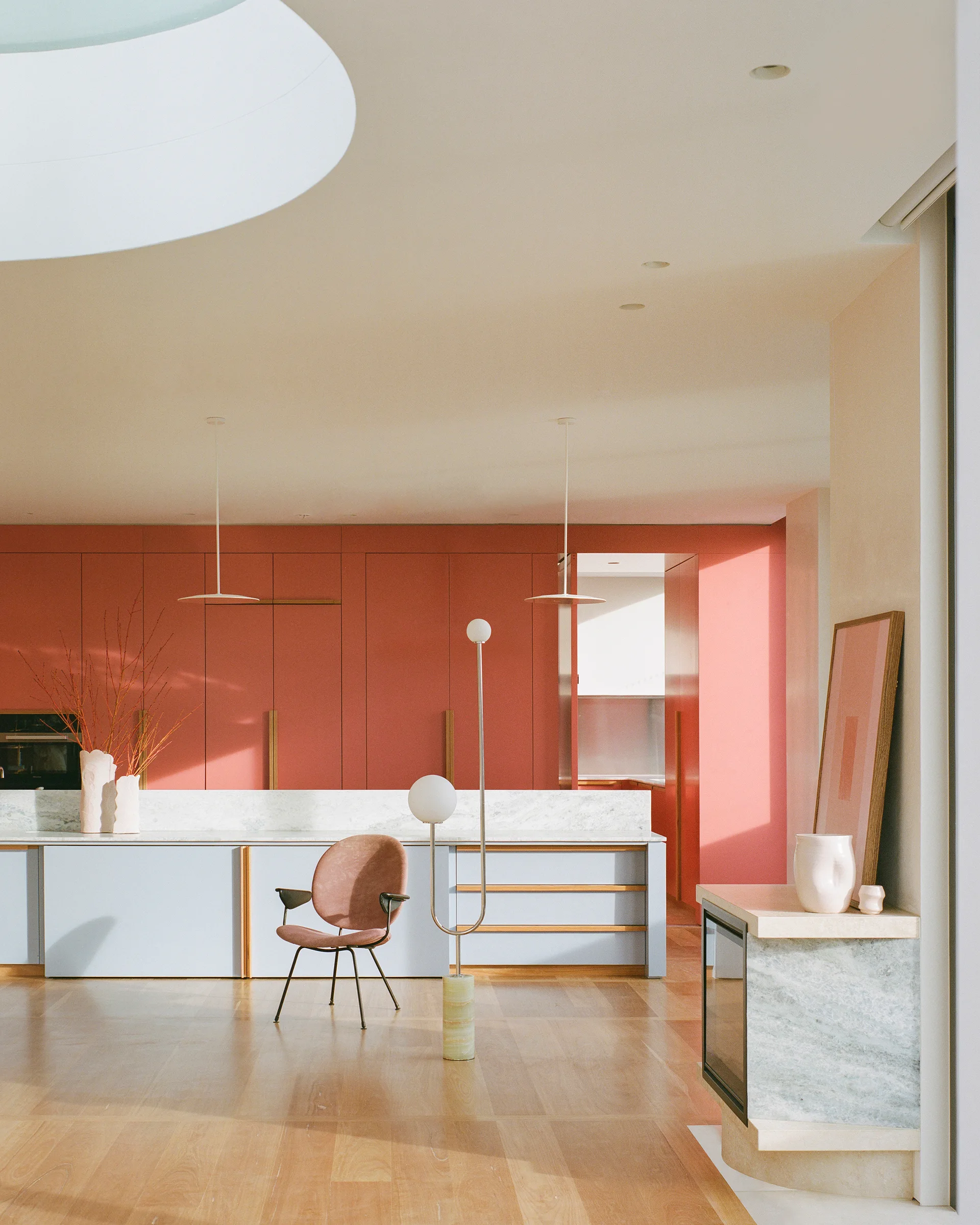 A modern kitchen with floor-to-ceiling coral cabinets, a light blue island with a marble countertop, and a sculptural floor lamp on light wood floors.