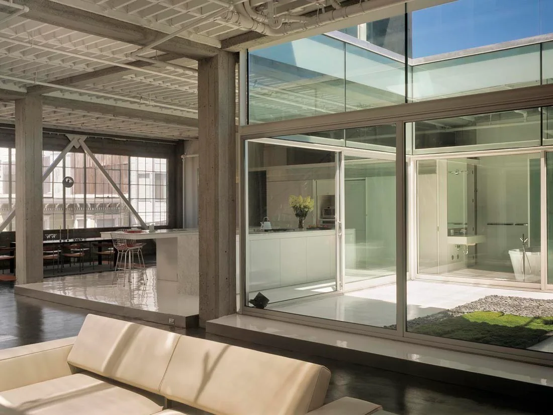 An open-plan loft featuring an interior glass courtyard, exposed concrete pillars, a cream leather sofa, and white painted pipes on the ceiling.