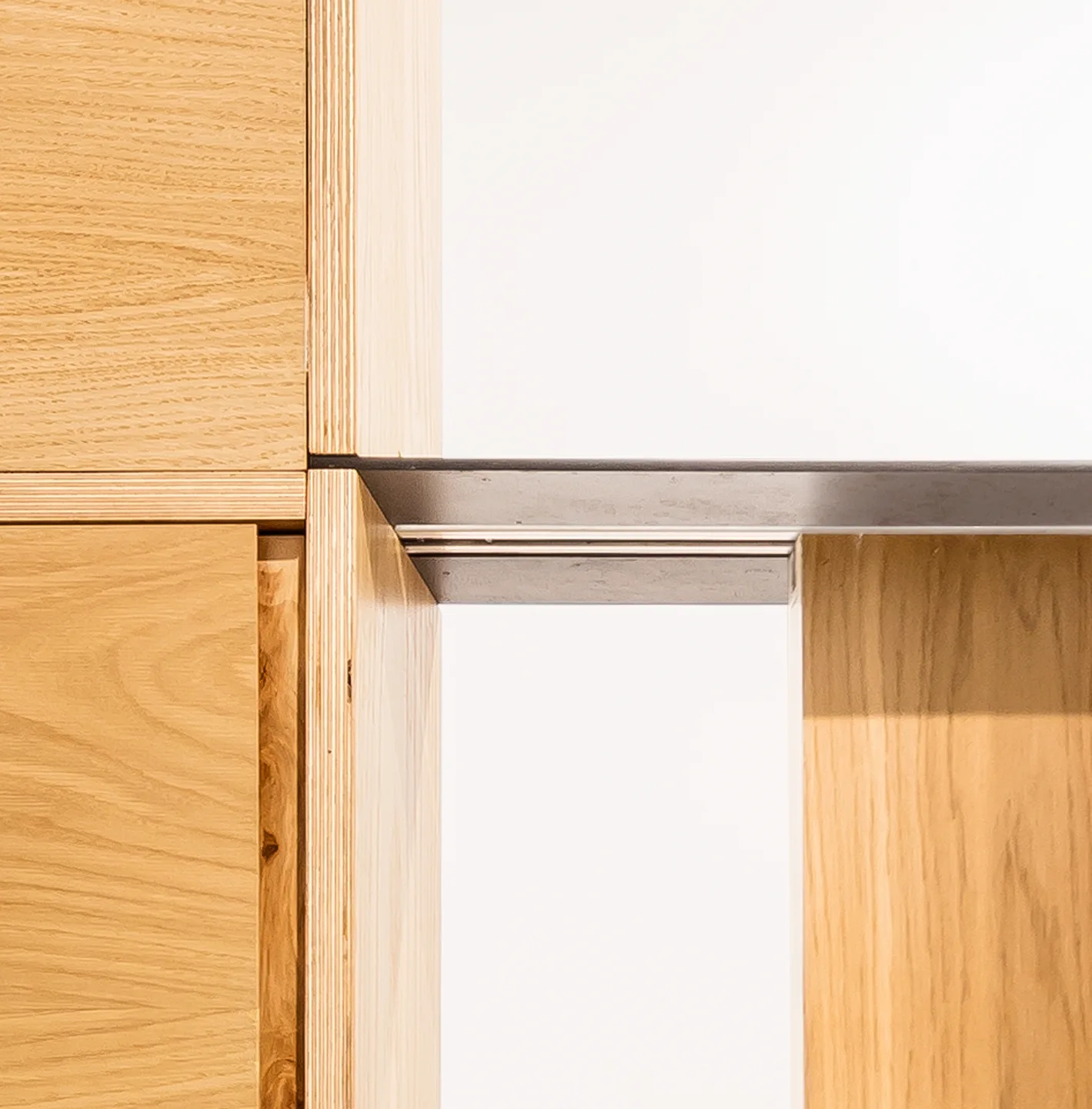 A close-up view of light wood cabinetry showcasing exposed plywood edges joined by a slim black metal support bar against a white background.