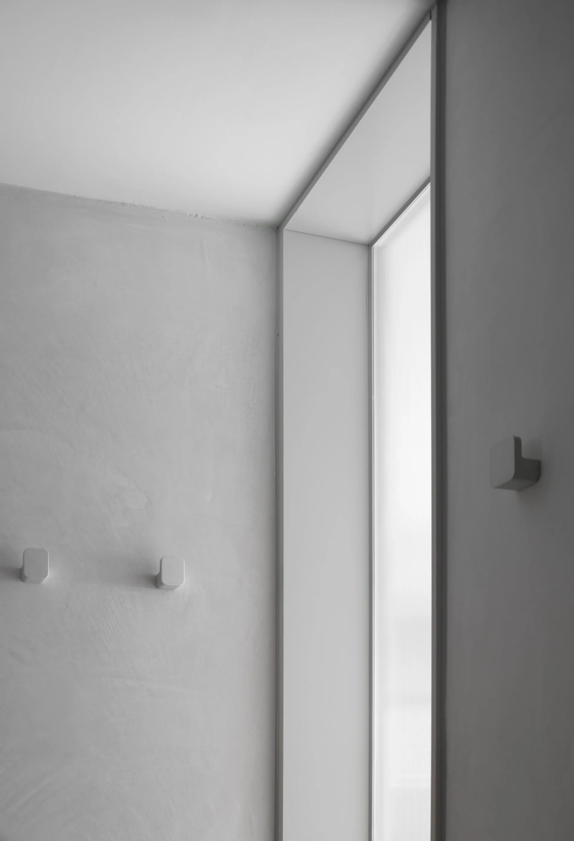 A smooth light grey plaster wall features three small square hooks next to a narrow vertical window casting soft diffuse light.