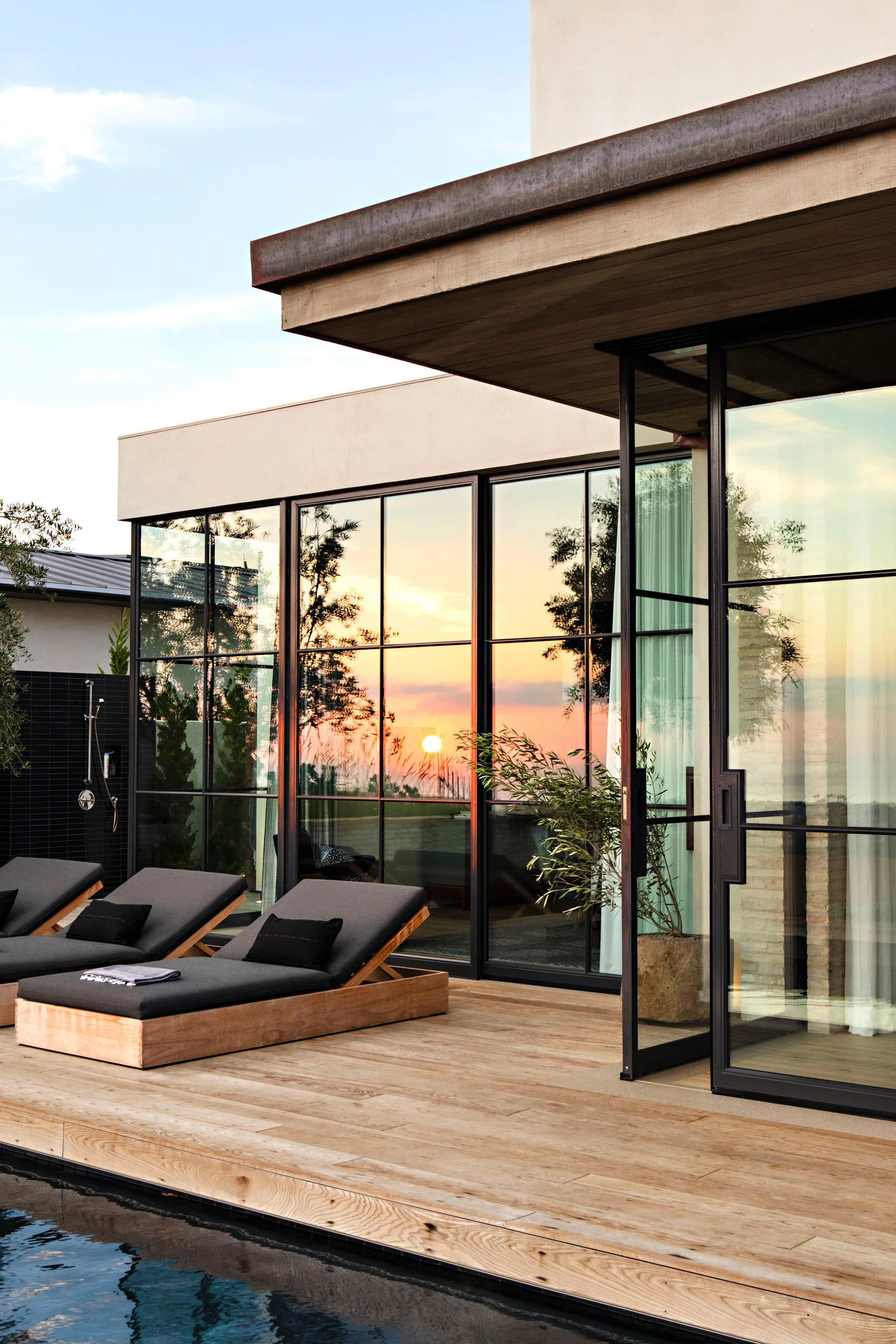 Charcoal-cushioned wooden loungers sit on a wood deck next to a pool, facing large black-framed grid glass windows reflecting a sunset.