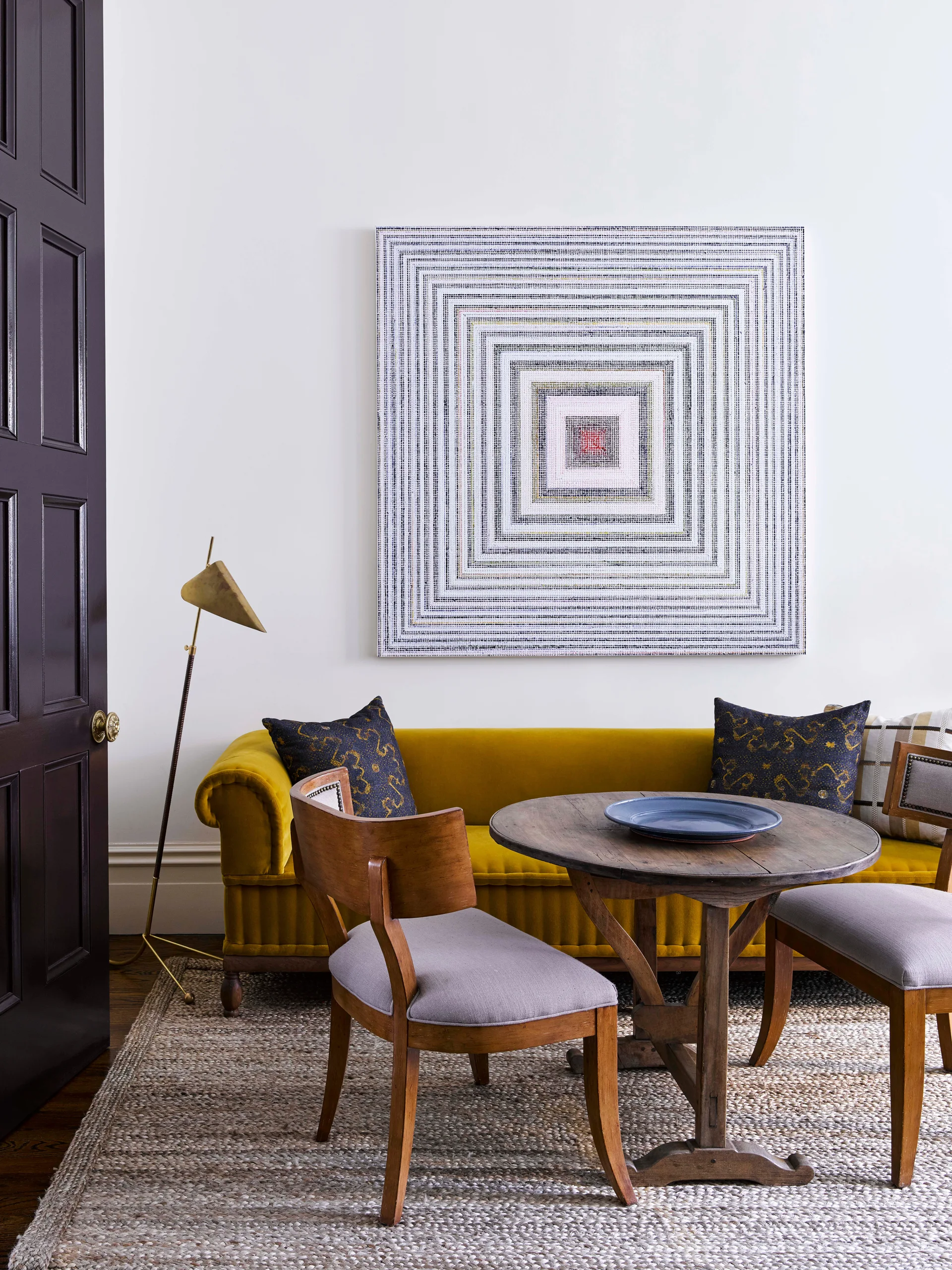 An ochre yellow velvet sofa sits behind a round wood dining table with upholstered chairs, under a large square piece of geometric wall art.