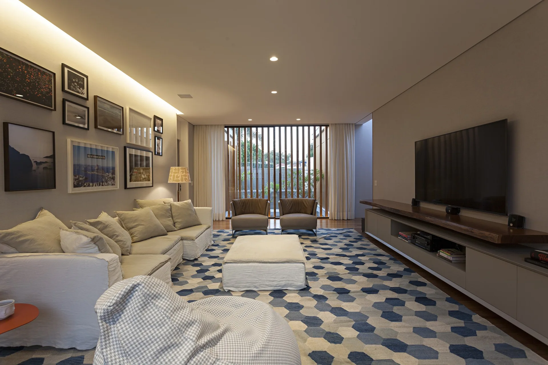 A contemporary living room features a cream sectional sofa, a large blue and beige hexagon pattern rug, a low media console, and a vertical wood slat screen.