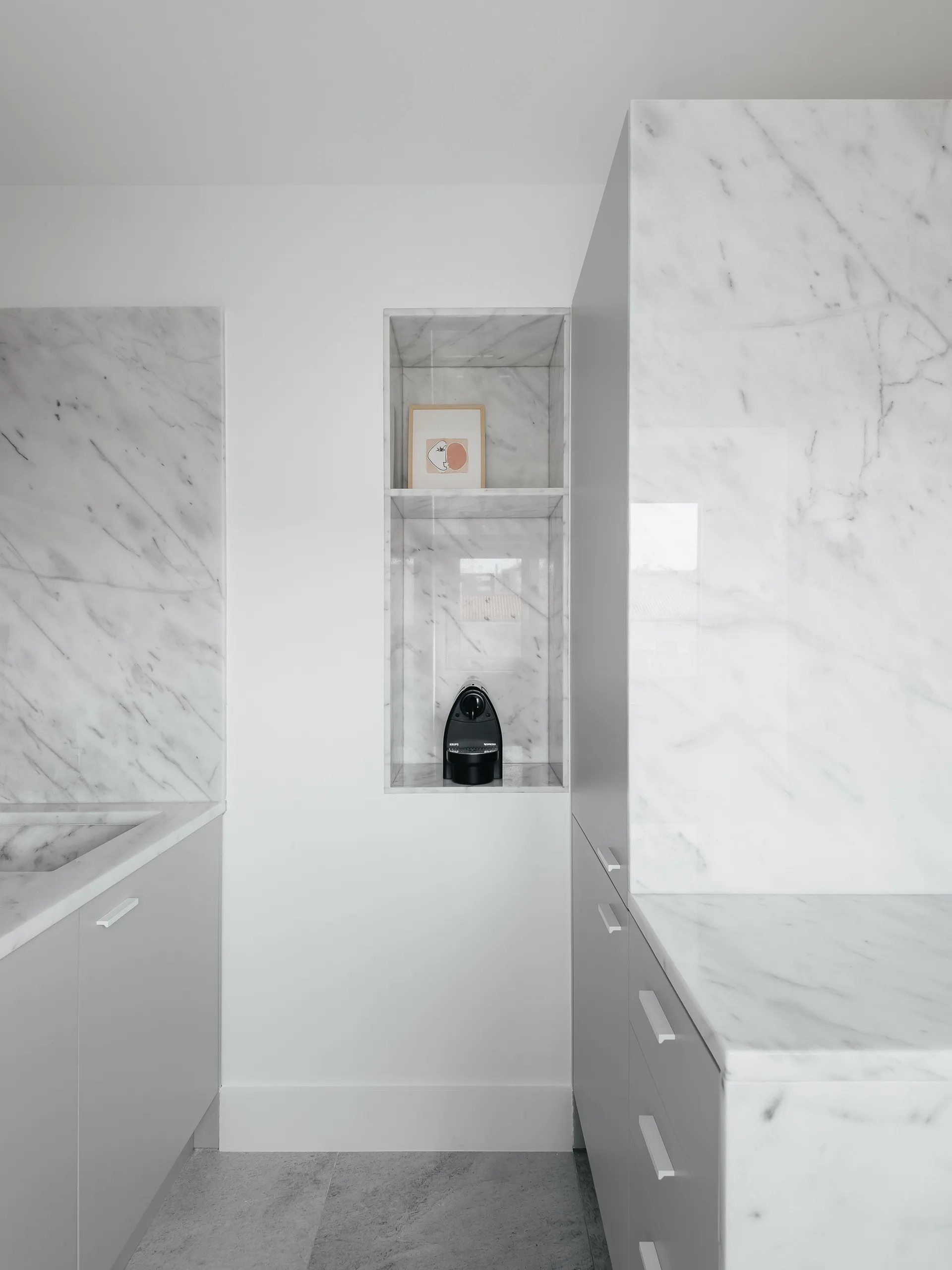 A minimalist kitchen features white cabinets, polished marble countertops, and a recessed marble wall niche holding a black espresso machine.