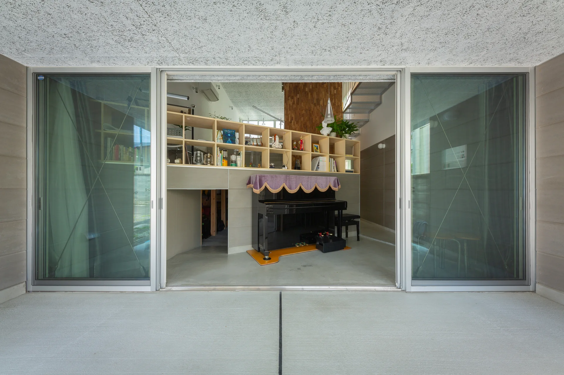 A view through open glass sliding doors reveals a minimalist interior with a black upright piano, a light wood grid bookshelf, and gray concrete flooring.