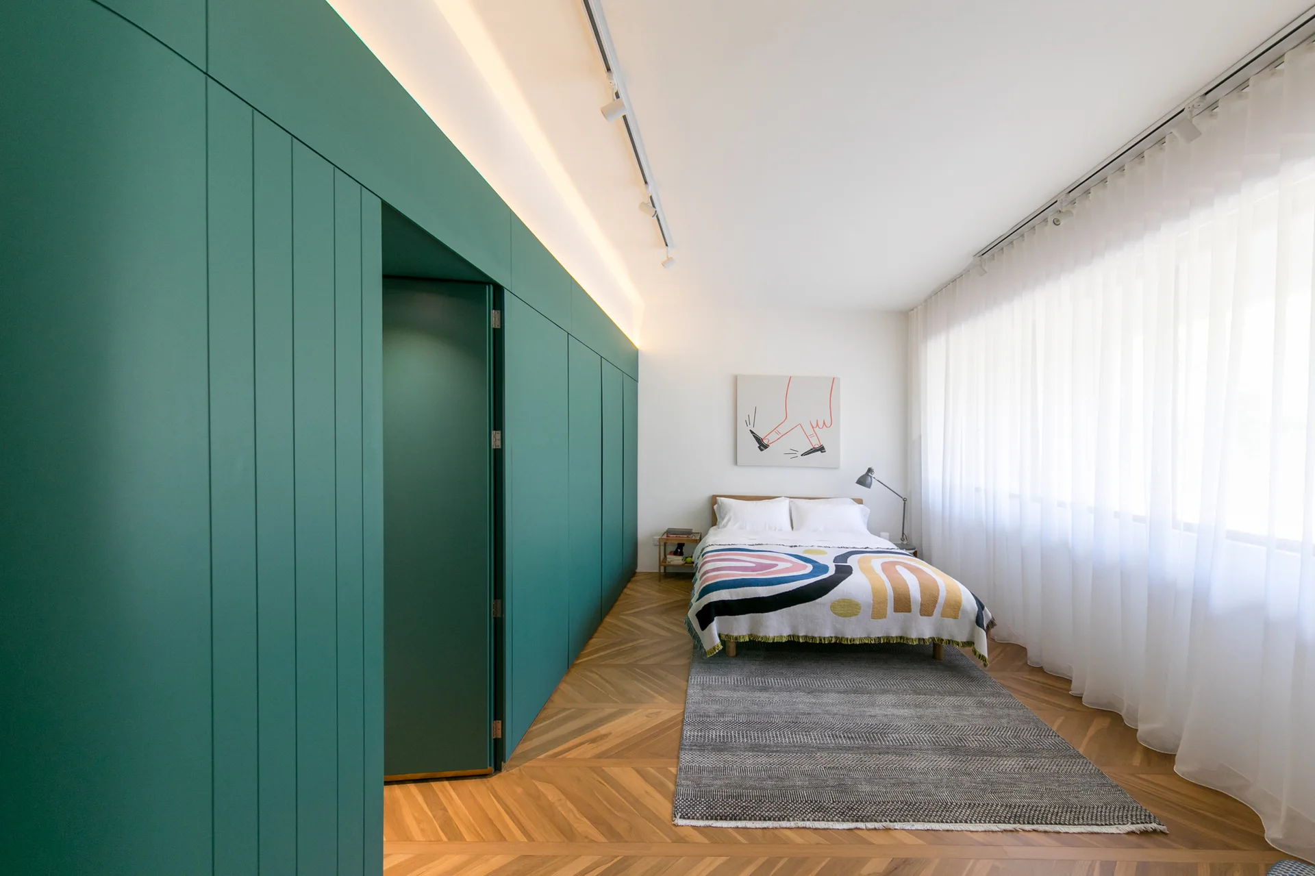 A modern bedroom features a bed with a patterned throw, herringbone wood flooring, and a large matte green cabinetry wall with integrated lighting.