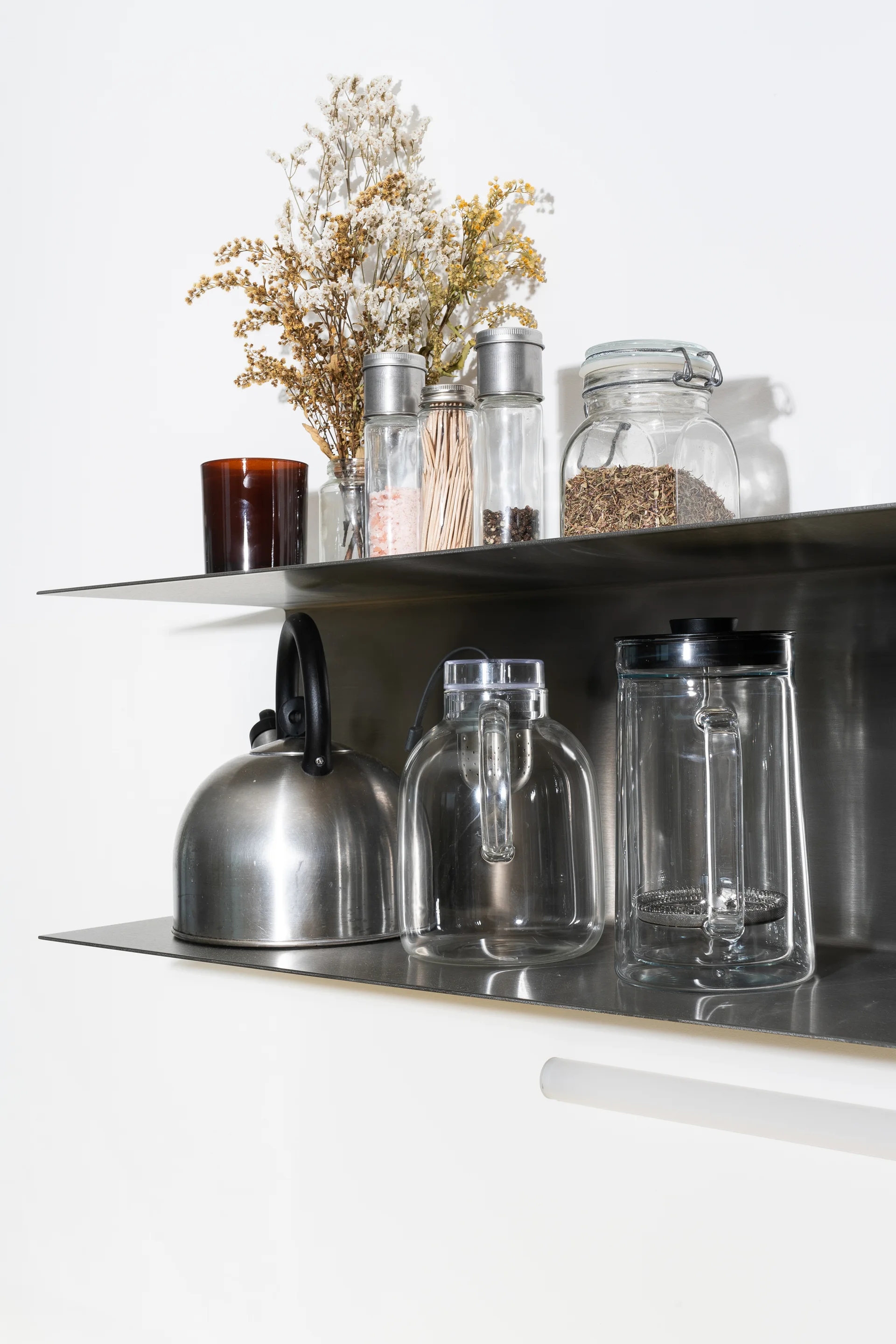 Two stainless steel wall shelves hold glass jars, a metal tea kettle, a french press, and dried flowers against a plain white wall.