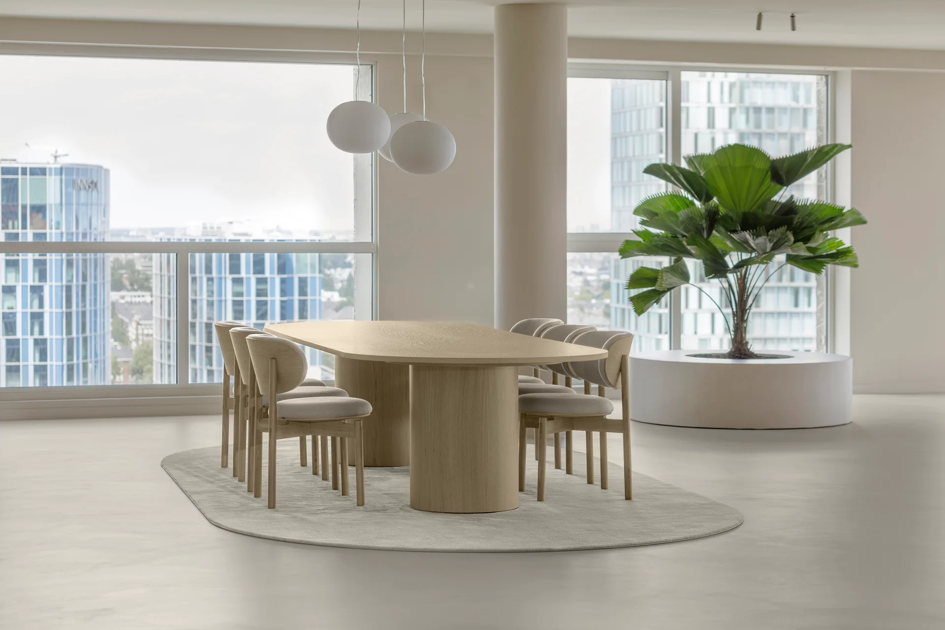 A minimalist dining room features a light wood oval table with chairs on an oval rug, near floor-to-ceiling windows and a large potted palm.